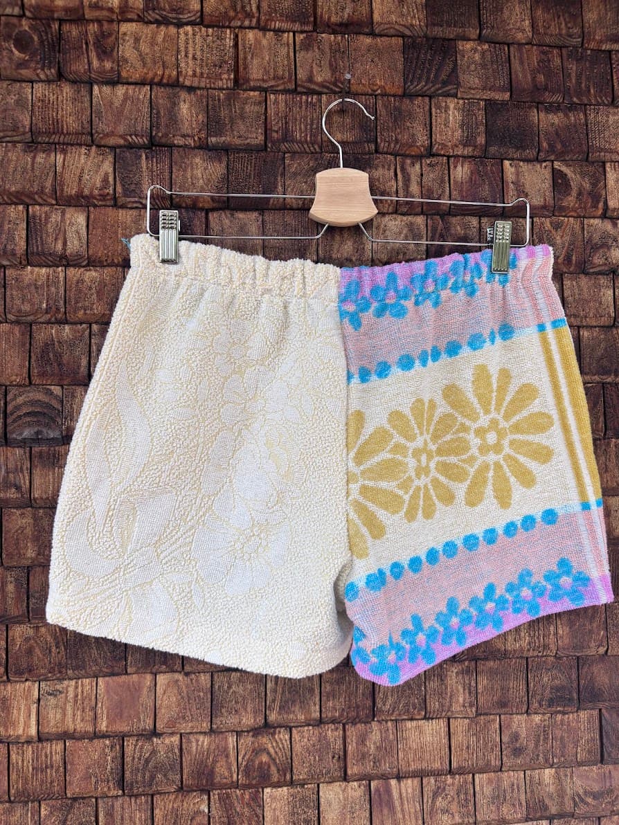 Rückansicht der handgefertigten Candy Riviera ‘80 Upcycling-Shorts, die das harmonische Patchwork-Design aus strukturierter cremeweißer Frottee-Seite und der verspielten, floral gemusterten Vintage-Hälfte zeigt, hängend vor einer rustikalen Holzwand.
