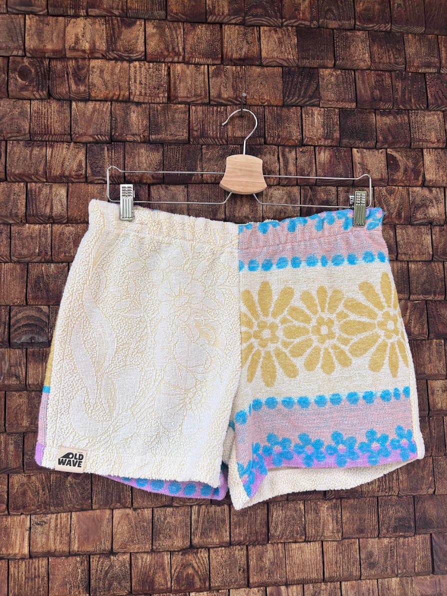 Vorderansicht der handgefertigten Candy Riviera ‘80 Shorts aus upgecycelten Vintage-Handtüchern. Eine Hälfte in schlichtem Creme-Weiß mit Frottee-Struktur, die andere Hälfte mit einem bunten Retro-Blumenmuster in Gelb, Hellblau und Pastell-Flieder, hängend vor einer Holzwand.
