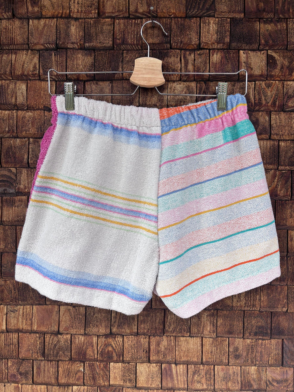 Rückansicht der handgefertigten Capri stripe '78 Upcycling-Shorts mit durchgehendem, horizontalem Streifenmuster in Pastellblau, Gelb und Rosa auf weißem Frottee-Stoff vor einer rustikalen Holzwand.
