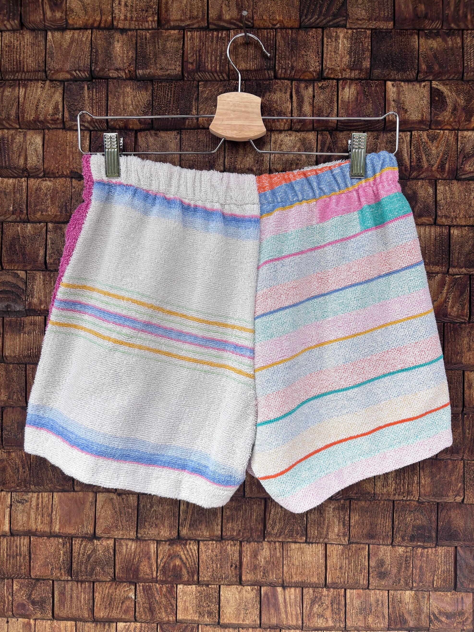 Rückansicht der handgefertigten Capri stripe '78 Upcycling-Shorts mit durchgehendem, horizontalem Streifenmuster in Pastellblau, Gelb und Rosa auf weißem Frottee-Stoff vor einer rustikalen Holzwand.
