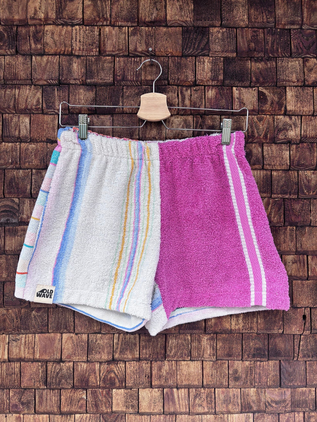 Vorderansicht einer handgefertigten Capri stripe '78 Shorts aus upgecycelten Vintage-Handtüchern. Rechte Seite im gestreiften Pastell-Design, linke Seite in kräftigem Pink-Beere, hängend vor einer Holzwand.