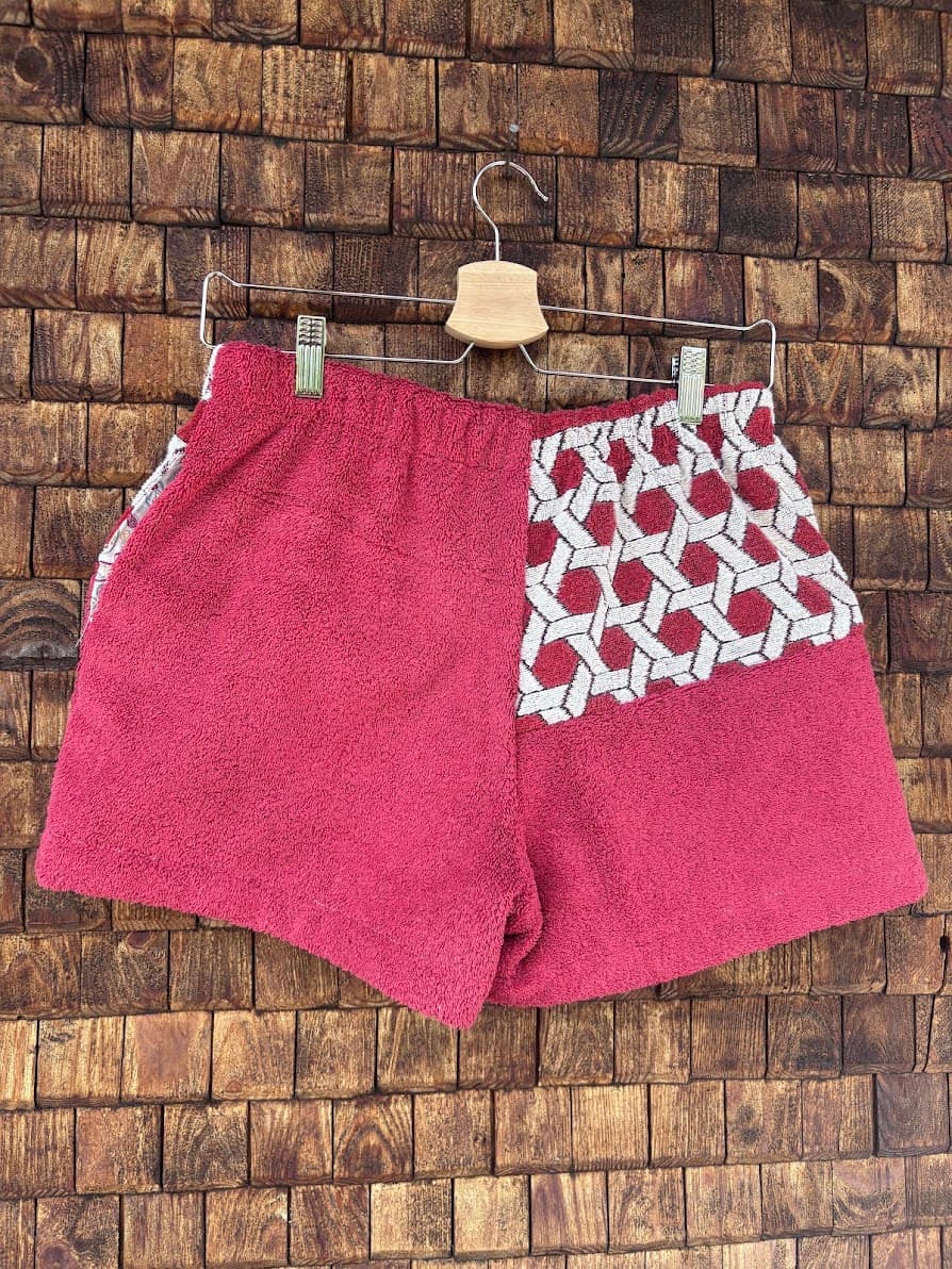 Rückansicht der Dusty mosaic '85 Upcycling-Shorts, die das einfarbige, strukturierte Frottee in Beerentönen und den kontrastreichen Mosaik-Stoffabschnitt zeigt, präsentiert vor einer rustikalen Holzwand.