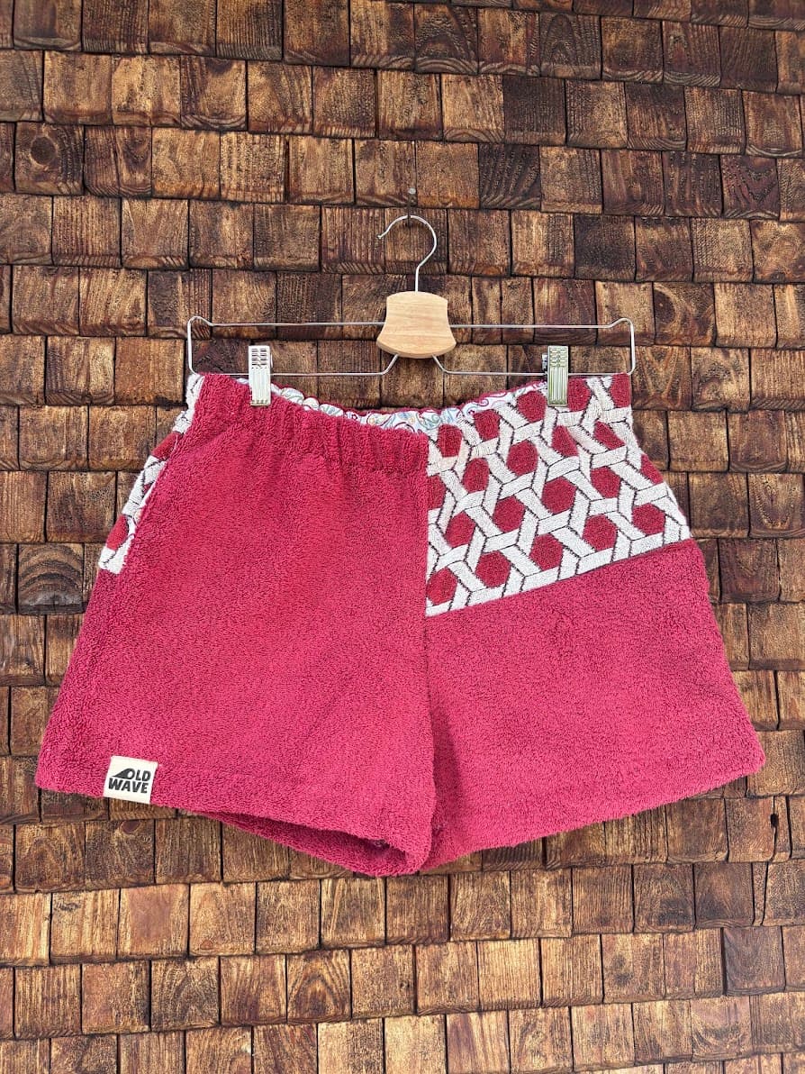 Vorderansicht der handgefertigten Dusty mosaic '85 Shorts aus upgecycelten Vintage-Handtüchern in kräftigem Magenta-Beere mit einem geometrischen Mosaik-Muster-Panel auf einer Seite, hängend vor einer Holzwand.