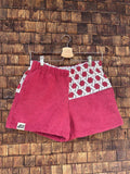 Vorderansicht der handgefertigten Dusty mosaic '85 Shorts aus upgecycelten Vintage-Handtüchern in kräftigem Magenta-Beere mit einem geometrischen Mosaik-Muster-Panel auf einer Seite, hängend vor einer Holzwand.