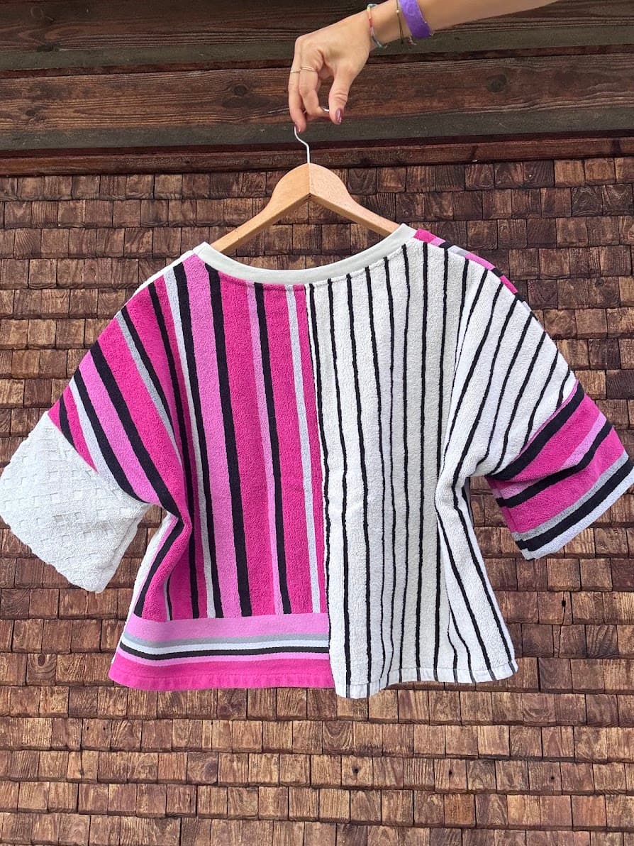 Rückansicht des Upcycling-Frotteeshirts „Strawberry '82“ von Old Wave an einem Kleiderbügel. Diese Perspektive zeigt das kontrastreiche Streifendesign auf der Rückseite: Der linke Ärmel und die linke Rückenhälfte führen das dicke, pink-schwarze Retro-Streifenmuster fort, während die rechte Hälfte in ein feineres, vertikales Streifenmuster aus schmalen schwarzen Linien auf weißem Grund übergeht. Der rechte Ärmel setzt sich in weißer Waffelstruktur ab.