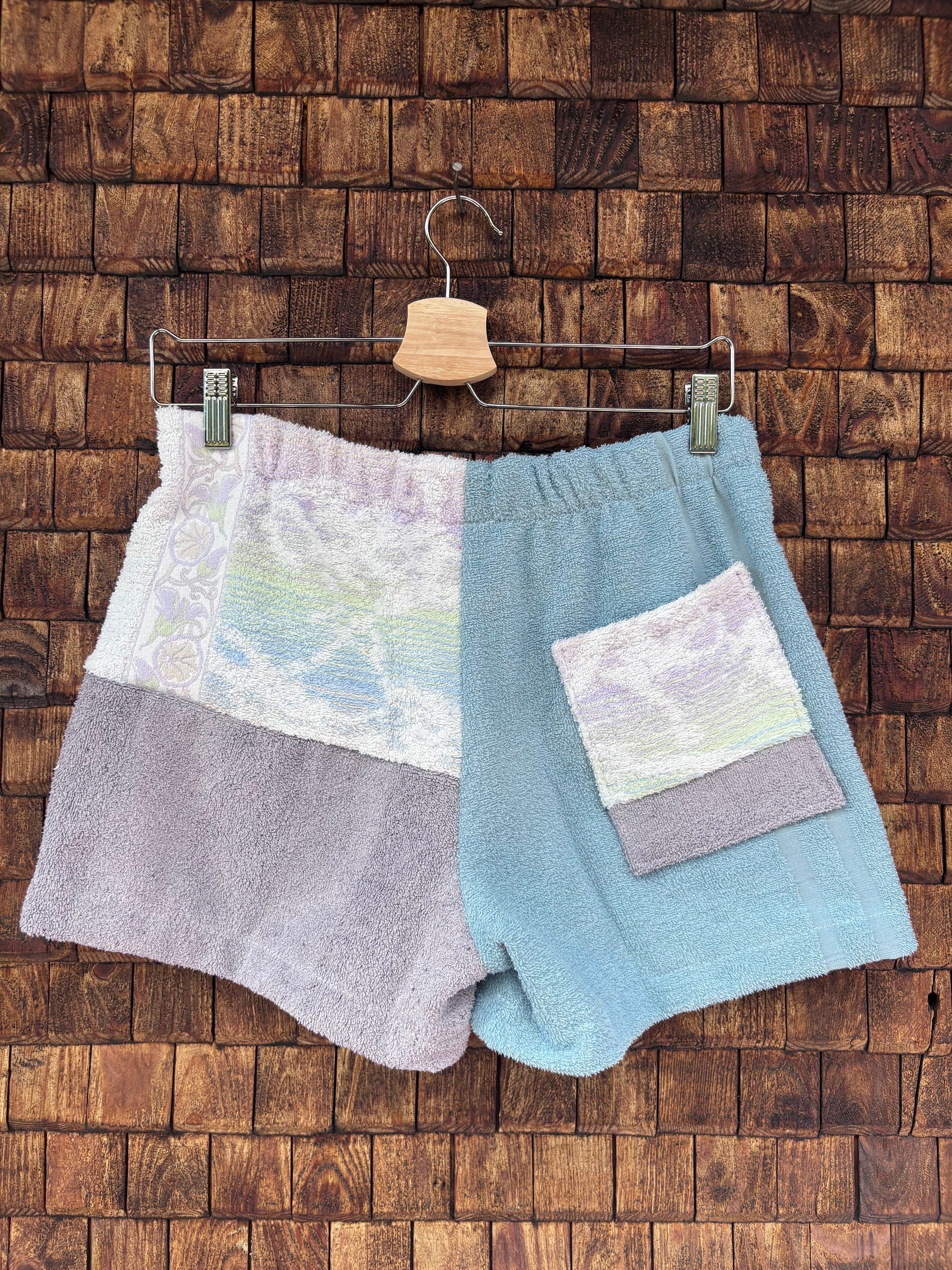 Rückansicht einer handgefertigten Mauve mosaic '67 Shorts aus upgecycelten Vintage-Handtüchern. Rechte Seite in softem Hellblau mit aufgesetzter Pastell-Gesäßtasche, linke Seite in Grau-Lila und gemustertem Weiß, hängend vor einer Holzwand.