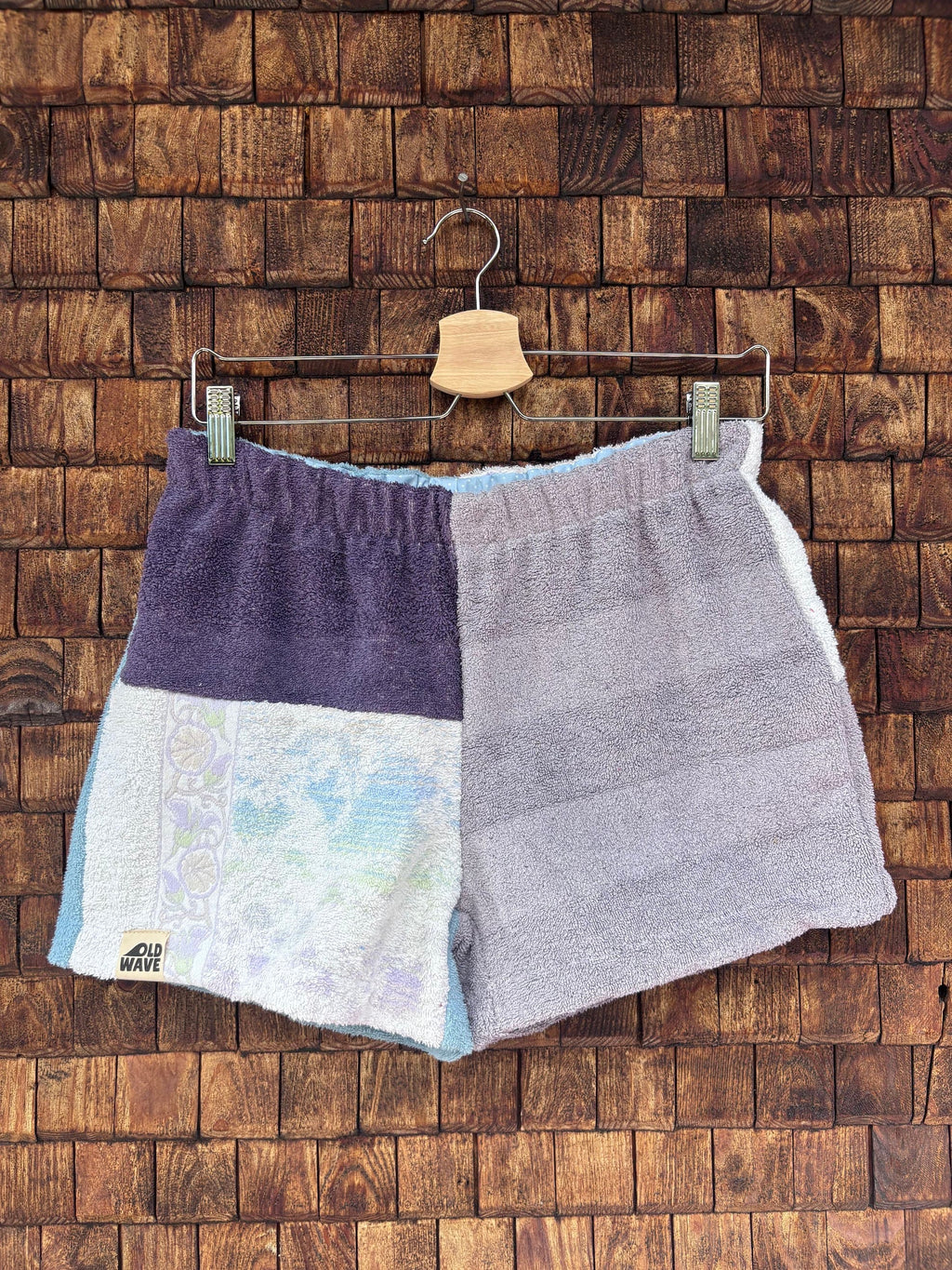 Vorderansicht der handgefertigten Mauve mosaic '67 Shorts im Patchwork-Design aus Frottee-Stoff in dunklem Mauve-Lila, softem Grau und hellen Abschnitten mit einem feinen, mosaikartigen Pastellmuster vor einer rustikalen Holzwand.