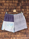 Vorderansicht der handgefertigten Mauve mosaic '67 Shorts im Patchwork-Design aus Frottee-Stoff in dunklem Mauve-Lila, softem Grau und hellen Abschnitten mit einem feinen, mosaikartigen Pastellmuster vor einer rustikalen Holzwand.