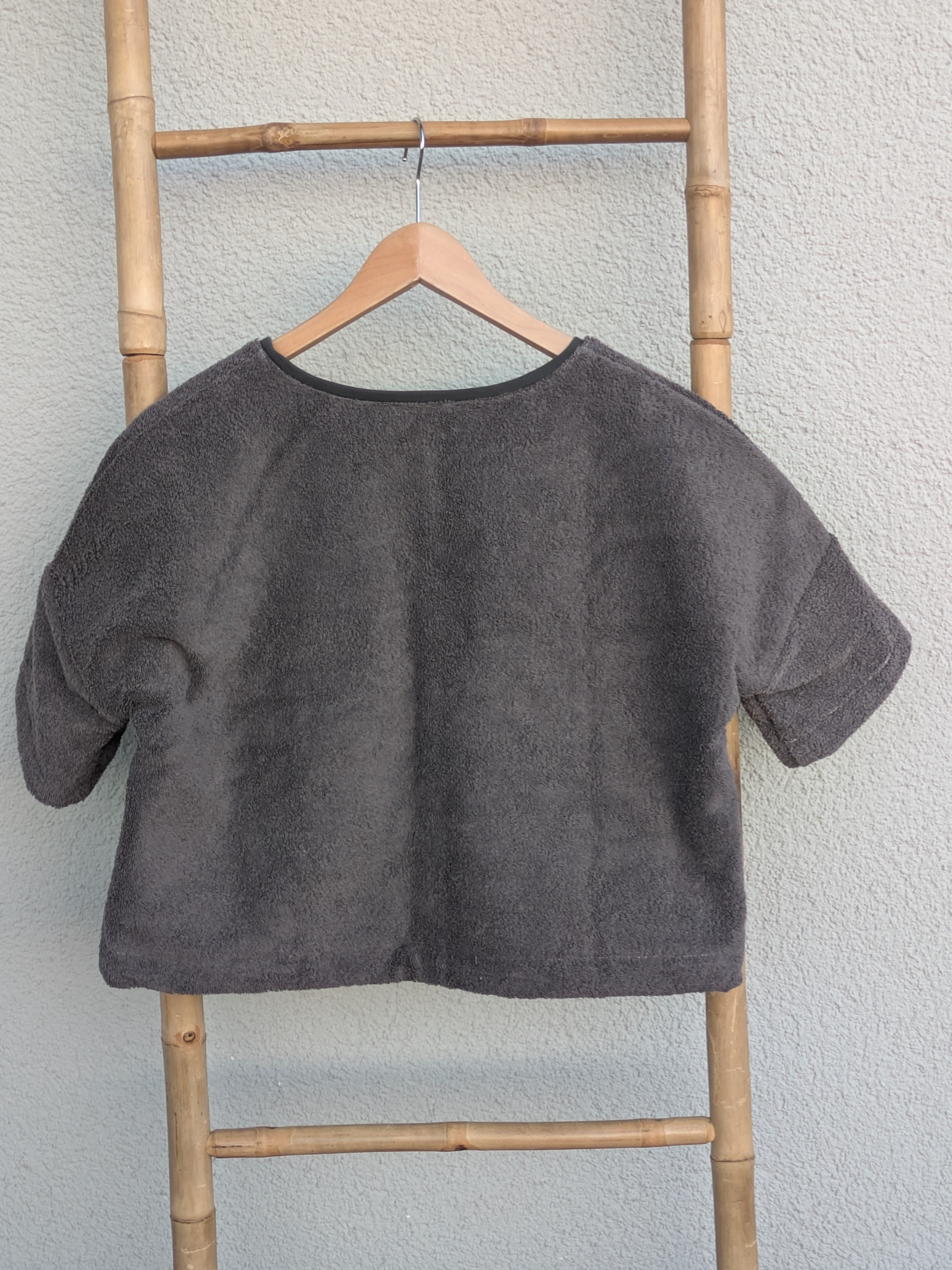 Rückansicht des dunkelgrauen Frottee Boxy Shirts 'Anthracite Wave ’71' von Old Wave. Zeigt den weiten Schnitt und die mittlere Naht. Das Shirt hängt an einem Holzkleiderbügel vor einer hellen Wand.