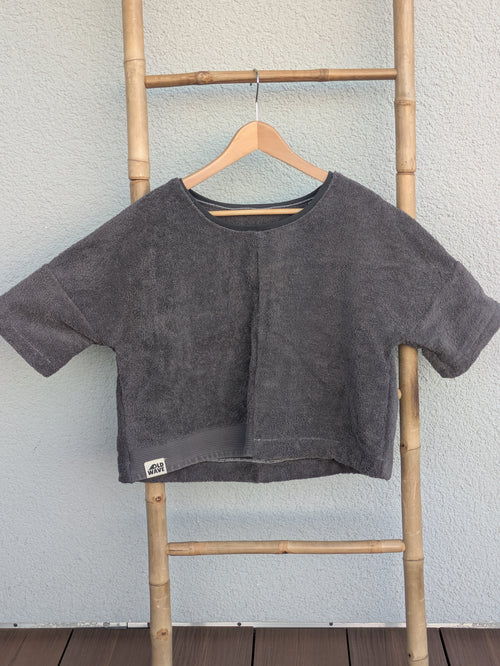 Vorderansicht des unifarbenen Frottee Boxy Shirts 'Anthracite Wave ’71' von Old Wave in Dunkelgrau. Mit Old Wave Logo-Patch unten links. Das Oberteil hängt auf einem Holzkleiderbügel an einer Bambusleiter.