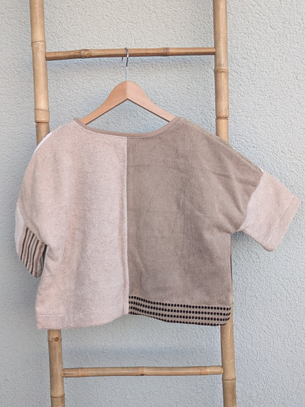 Rückansicht des Boxy Shirts 'Beige Coast ’70' von Old Wave im Half-and-Half-Look. Zeigt zwei unterschiedliche Beigetöne auf der linken und rechten Seite. Das Shirt hängt an einem Holzkleiderbügel vor einer hellen Wand.