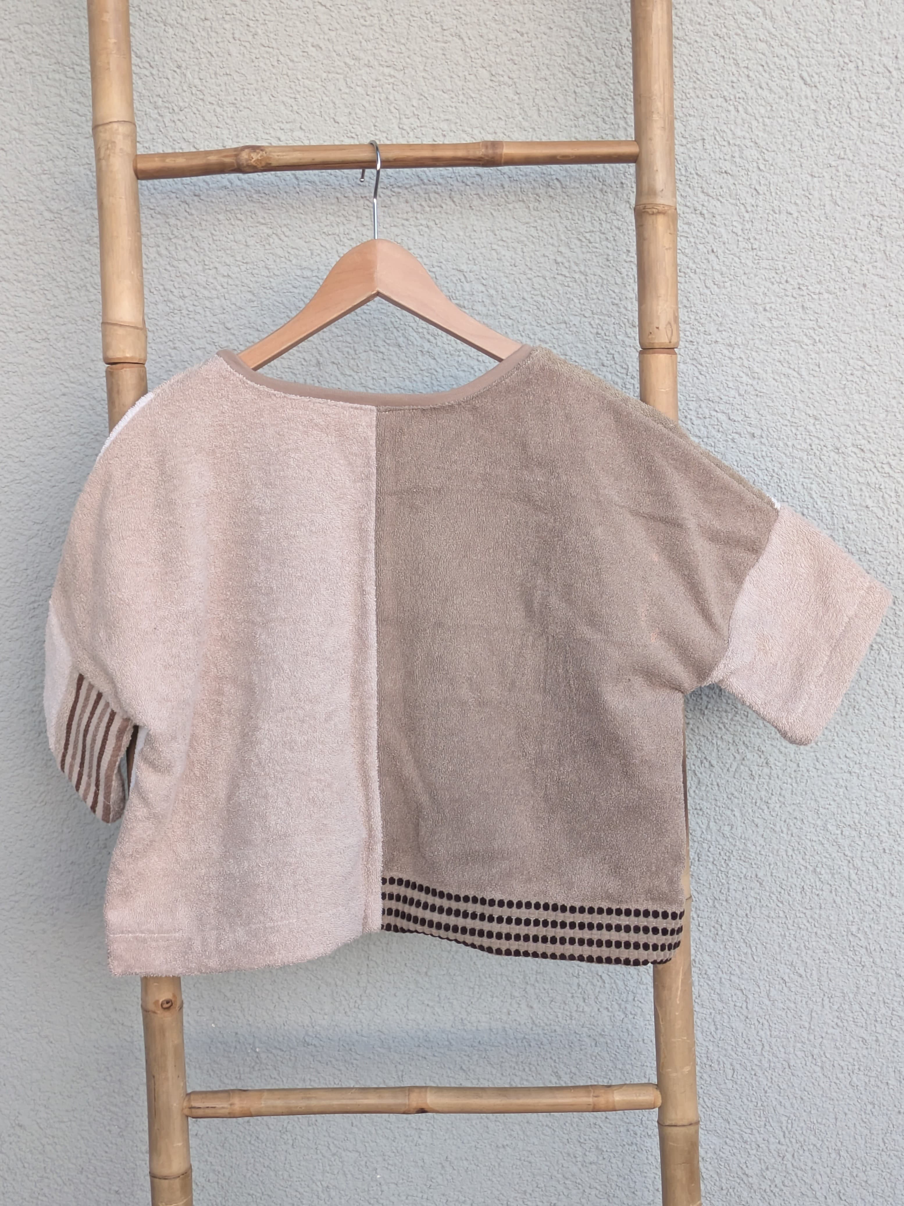Rückansicht des Boxy Shirts 'Beige Coast ’70' von Old Wave im Half-and-Half-Look. Zeigt zwei unterschiedliche Beigetöne auf der linken und rechten Seite. Das Shirt hängt an einem Holzkleiderbügel vor einer hellen Wand.