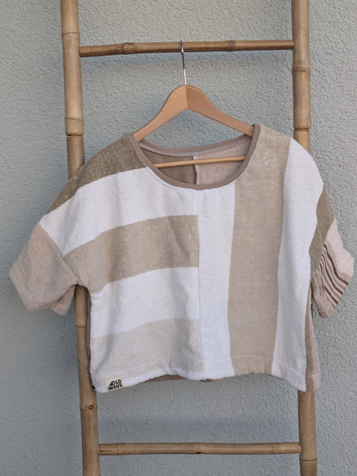 Vorderansicht des Frottee Boxy Shirts 'Beige Coast ’70' von Old Wave. Weit geschnittenes Oberteil aus Frottee mit sandfarbenen und weißen Blockstreifen. Das Shirt hängt auf einem Holzkleiderbügel an einer Bambusleiter.