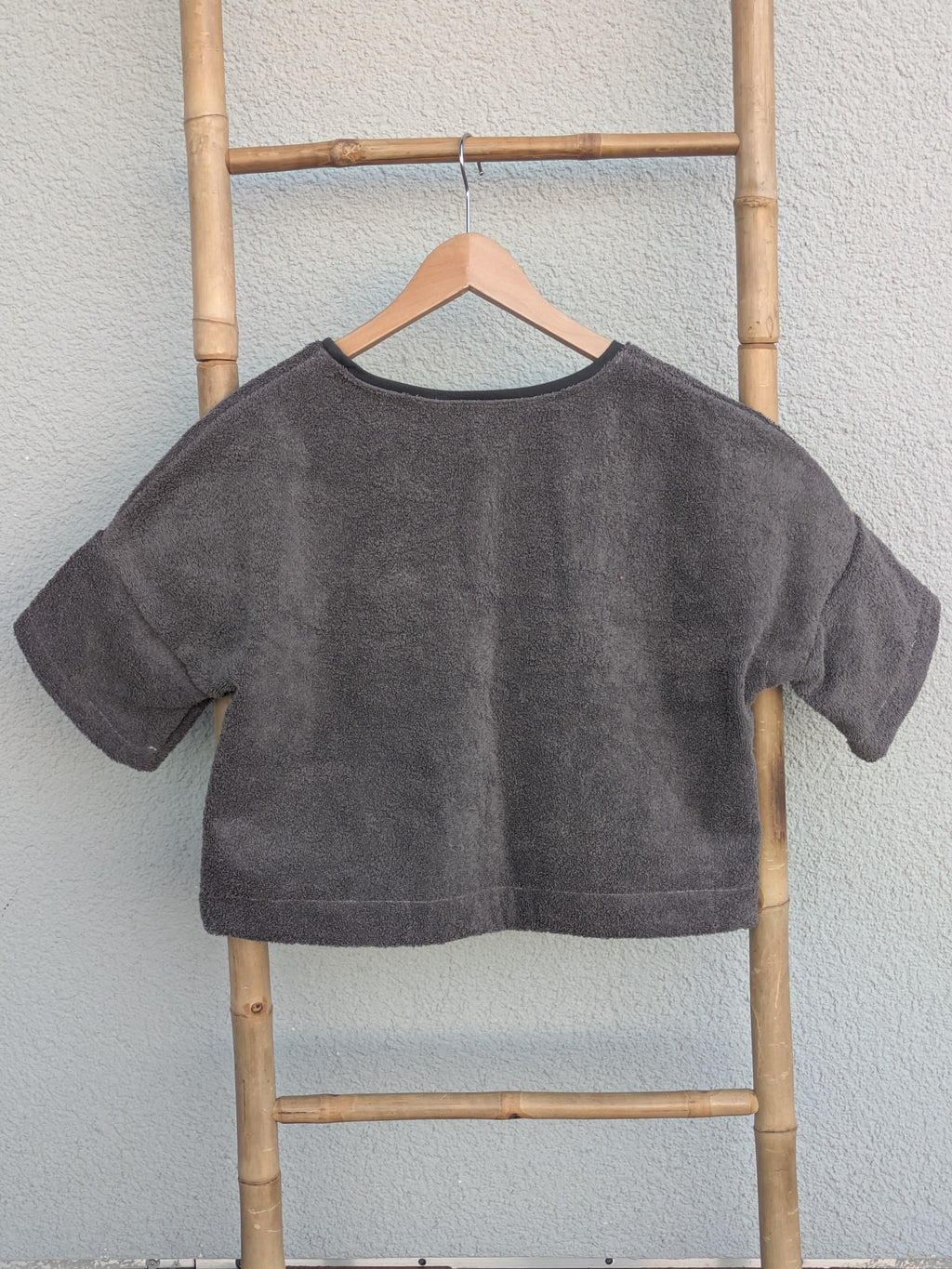 Rückansicht des dunkelgrauen Boxy Shirts 'Coal Coast ’73' von Old Wave. Zeigt den weiten Schnitt und den durchgehenden dunklen Frotteestoff. Das Shirt hängt an einem Kleiderbügel an einer Bambusleiter.