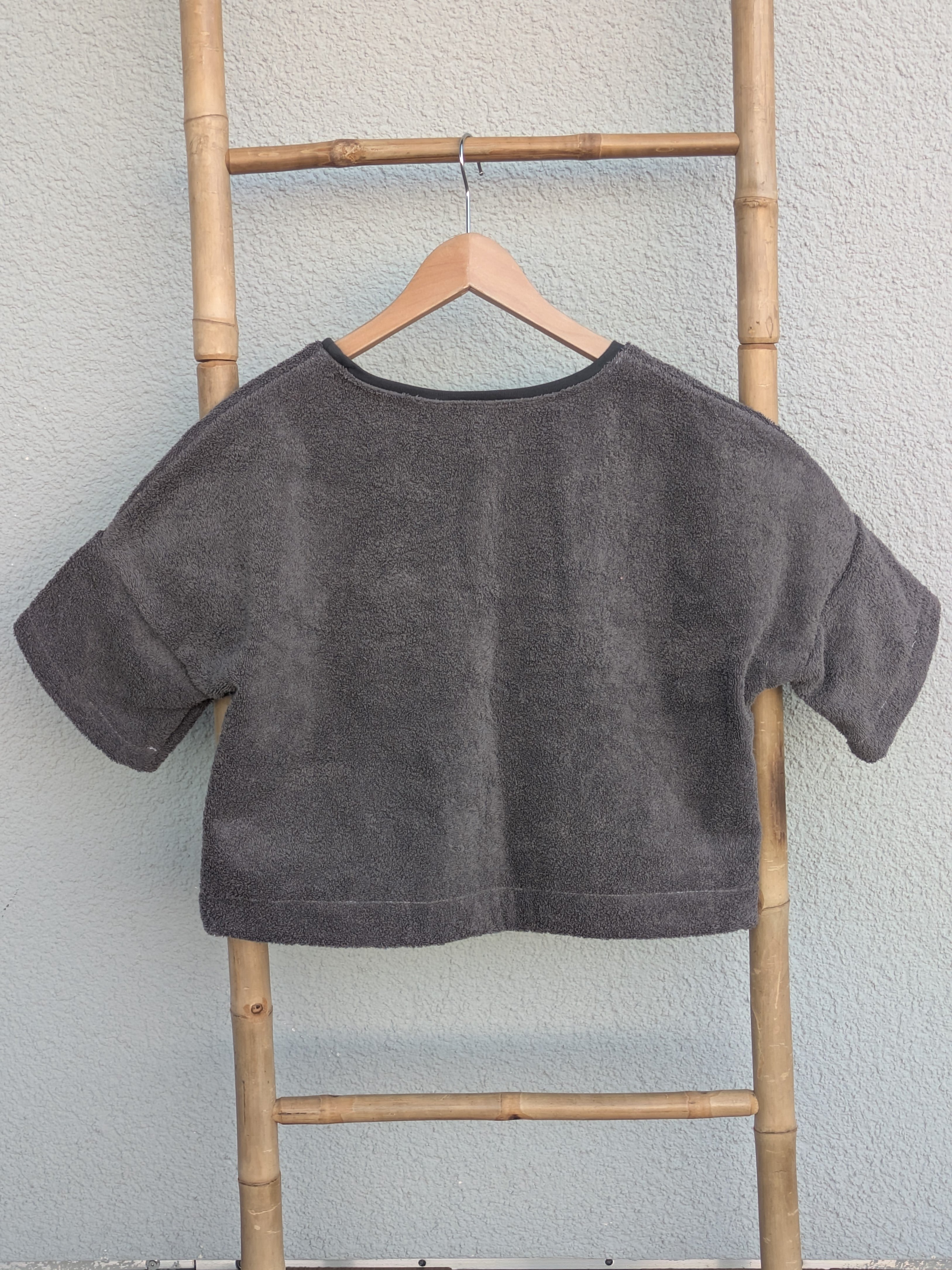 Rückansicht des dunkelgrauen Boxy Shirts 'Coal Coast ’73' von Old Wave. Zeigt den weiten Schnitt und den durchgehenden dunklen Frotteestoff. Das Shirt hängt an einem Kleiderbügel an einer Bambusleiter.