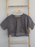 Vorderansicht des dunkelgrauen Frottee Boxy Shirts 'Coal Coast ’73' von Old Wave mit farblich abgesetztem Saumbund und Logo-Patch unten links. Das Shirt hängt an einem Holzkleiderbügel vor einer hellen Wand.