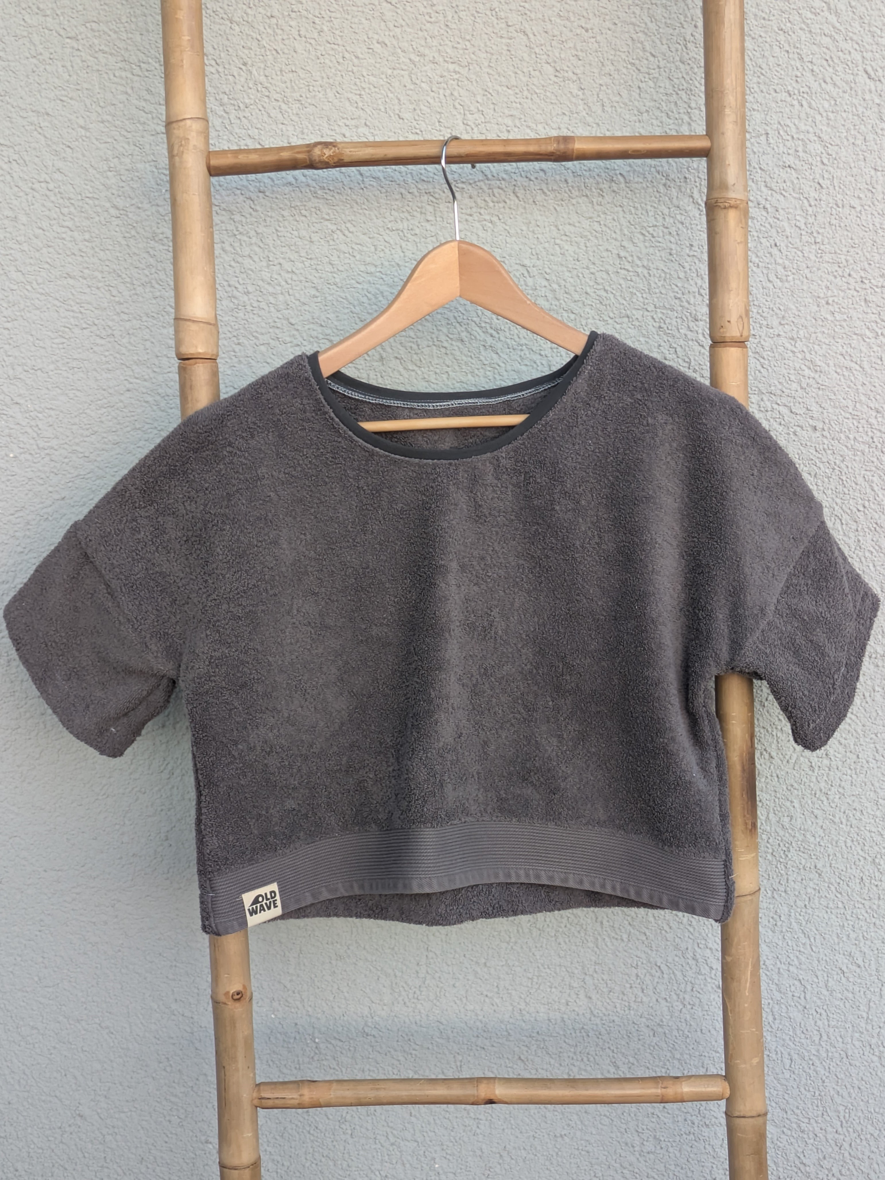 Vorderansicht des dunkelgrauen Frottee Boxy Shirts 'Coal Coast ’73' von Old Wave mit farblich abgesetztem Saumbund und Logo-Patch unten links. Das Shirt hängt an einem Holzkleiderbügel vor einer hellen Wand.