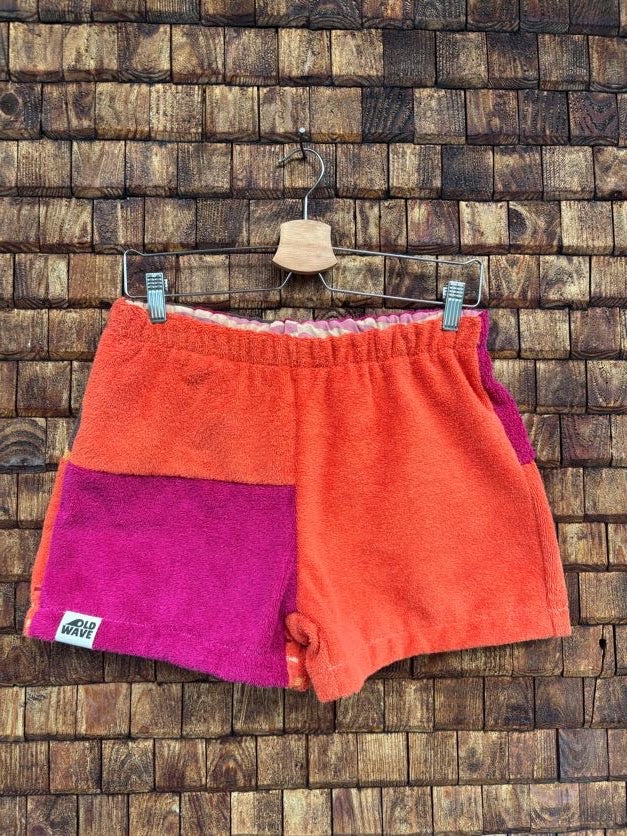 Vorderansicht der Frottee-Shorts 'Blueberry' von Old Wave in leuchtendem Orange mit einem magentafarbigen Akzent, elastischem Gummibund und Old Wave Logo-Patch vor einer Holzziegel-Wand.