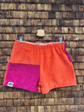 Vorderansicht der Frottee-Shorts 'Blueberry' von Old Wave in leuchtendem Orange mit einem magentafarbigen Akzent, elastischem Gummibund und Old Wave Logo-Patch vor einer Holzziegel-Wand.