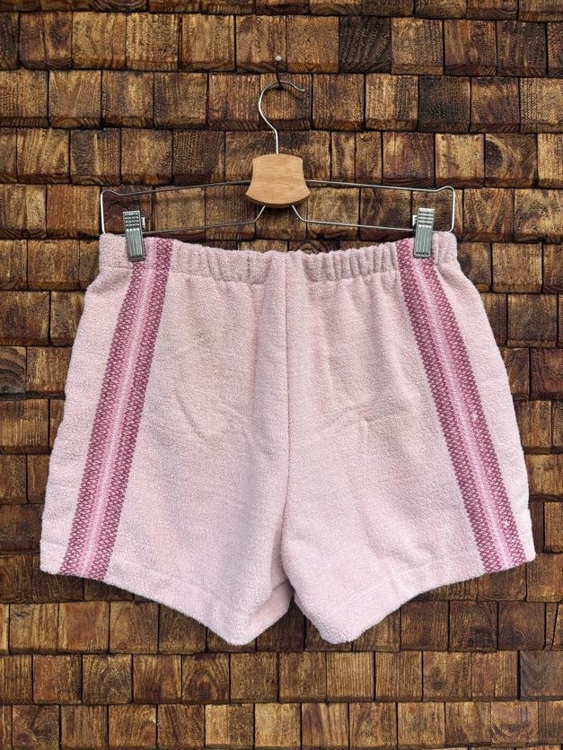 Rückansicht der Frottee-Shorts 'Rose-Shell' von Old Wave in hellem Rosé mit seitlich verlaufenden, altrosafarbenen Zierstreifen-Bordüren und elastischem Gummibund auf einem Kleiderbügel vor einer Holzziegel-Wand.