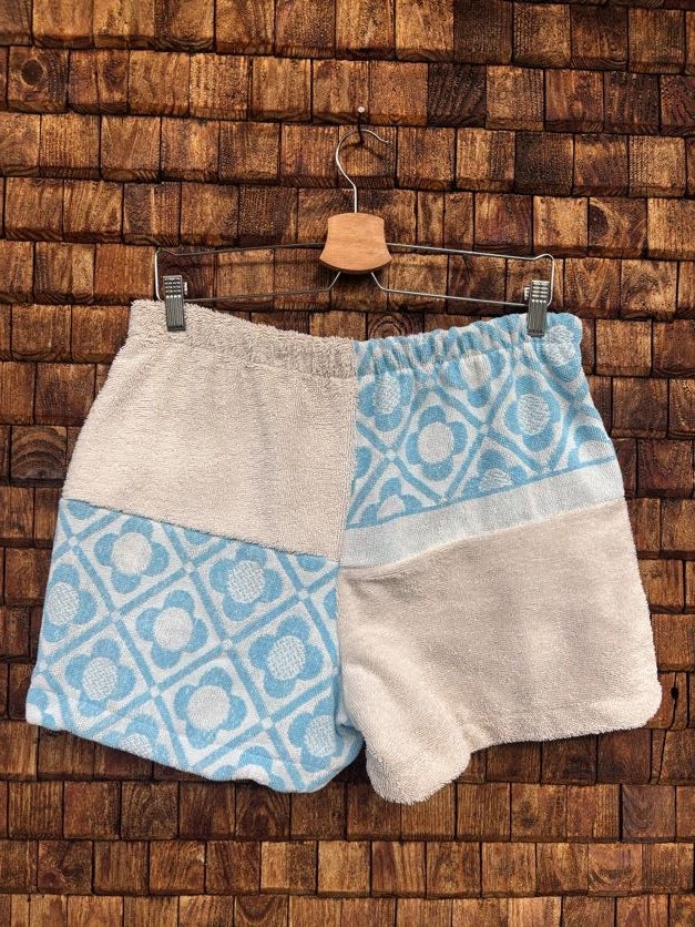 Rückansicht der Frottee-Shorts 'Shell' von Old Wave. Zeigt das viergeteilte Patchwork-Muster aus hellem Creme-Frottee und hellblau-weißen geometrischen Kachel-Ornamenten mit elastischem Gummibund auf einem Kleiderbügel.