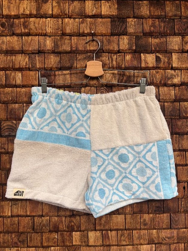 Vorderansicht der Frottee-Shorts 'Shell' von Old Wave im abwechslungsreichen Patchwork-Design aus naturweißem Frottee und blau-weißem Retro-Ornamentmuster mit Old Wave Logo-Patch unten rechts vor einer Holzziegel-Wand.