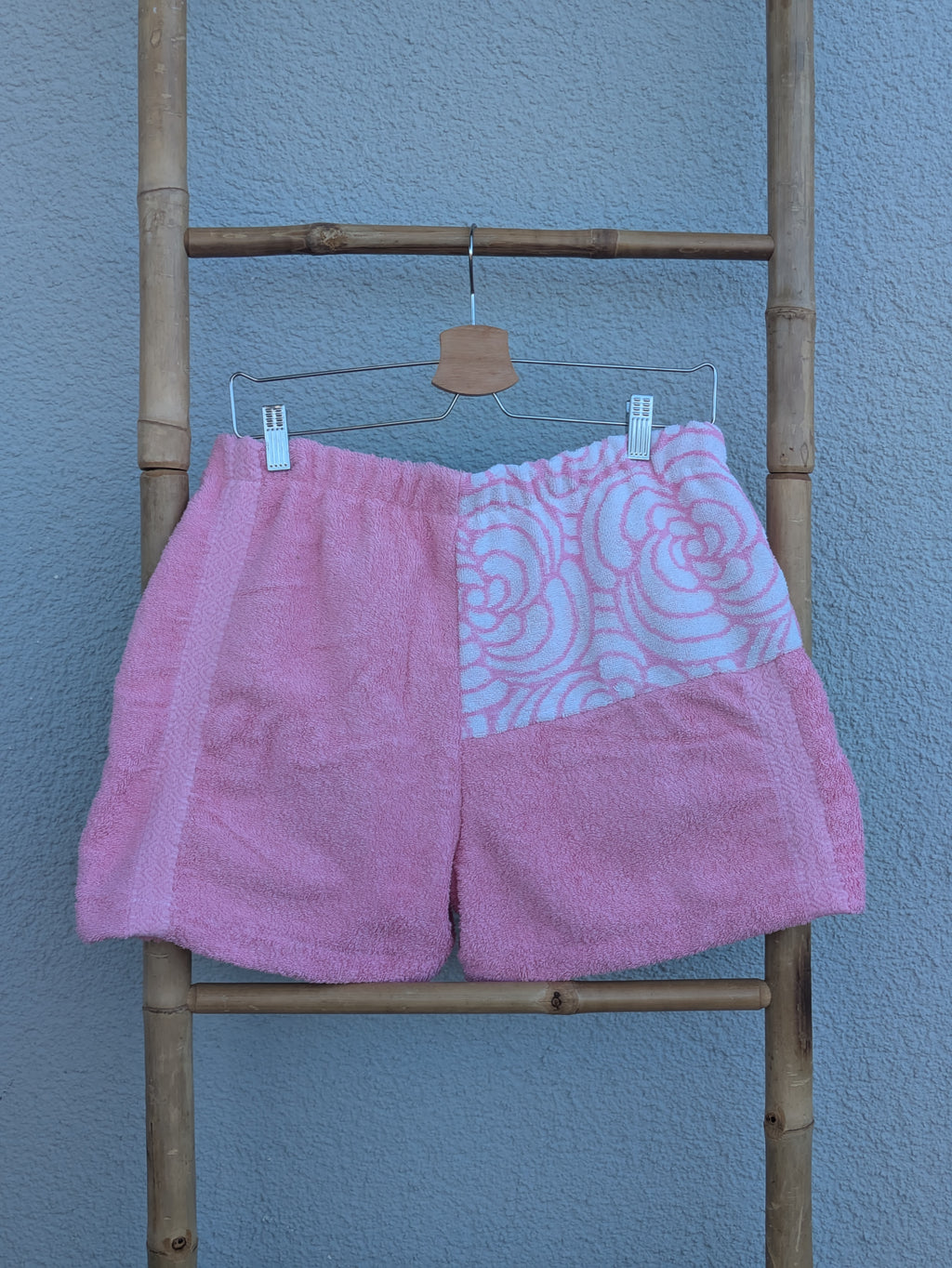 Rückansicht der Frottee Towel Shorts 'Bubblegum Retro ’73' von Old Wave. Zeigt das überwiegend unifarben rosa Design mit einem gemusterten Passen-Einsatz oben rechts vor einer hellen Wand.