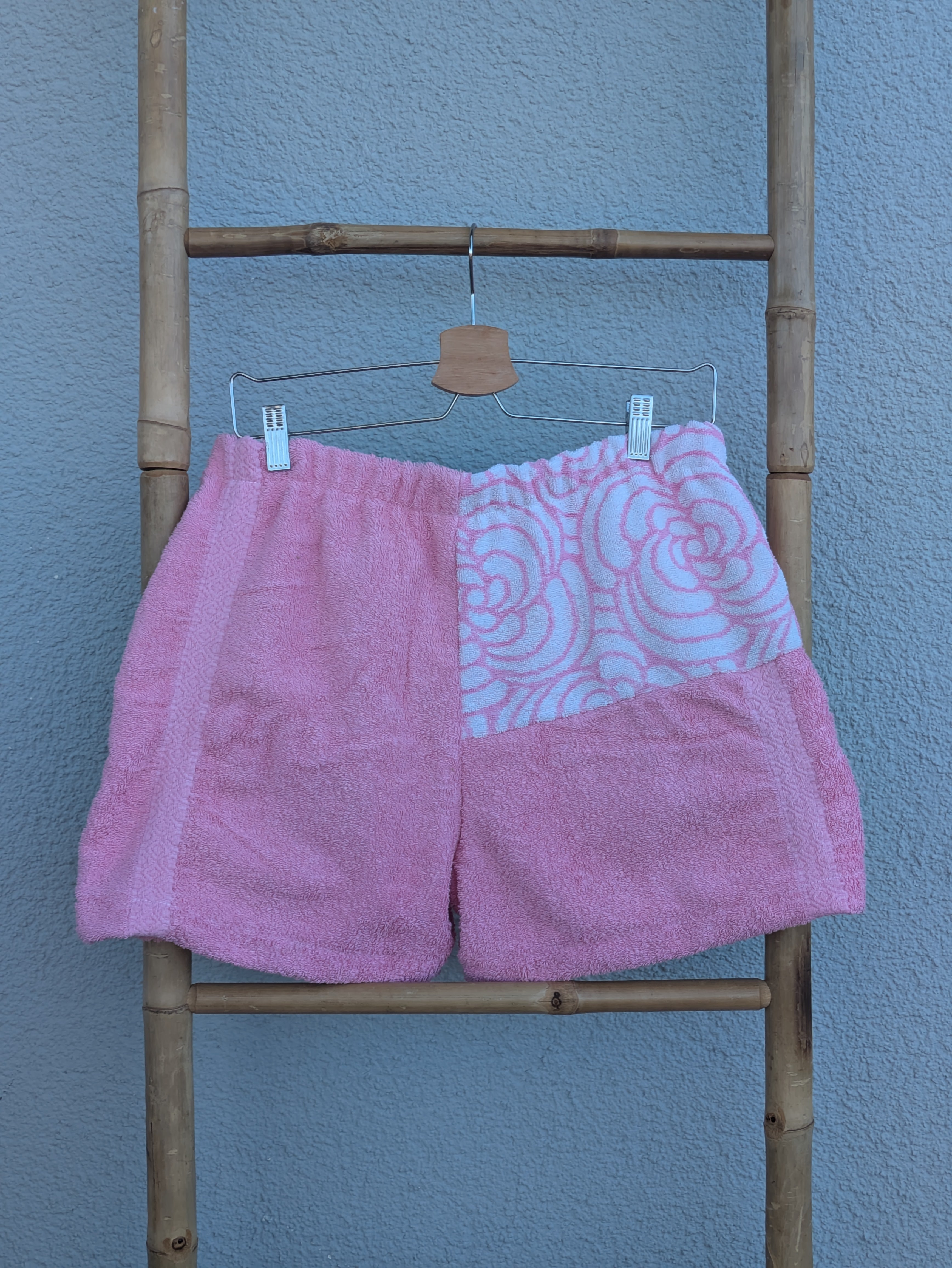 Rückansicht der Frottee Towel Shorts 'Bubblegum Retro ’73' von Old Wave. Zeigt das überwiegend unifarben rosa Design mit einem gemusterten Passen-Einsatz oben rechts vor einer hellen Wand.
