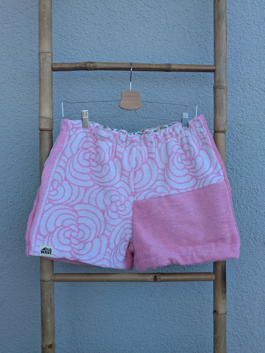 Vorderansicht der Frottee Towel Shorts 'Bubblegum Retro ’73' von Old Wave in Pastellrosa und Weiß. Patchwork-Design mit weißem, rosa gemustertem Wellen- und Bogen-Frottee oben und einem unifarbenen rosa Hosenbein-Einsatz unten rechts, präsentiert an einer Bambusleiter.