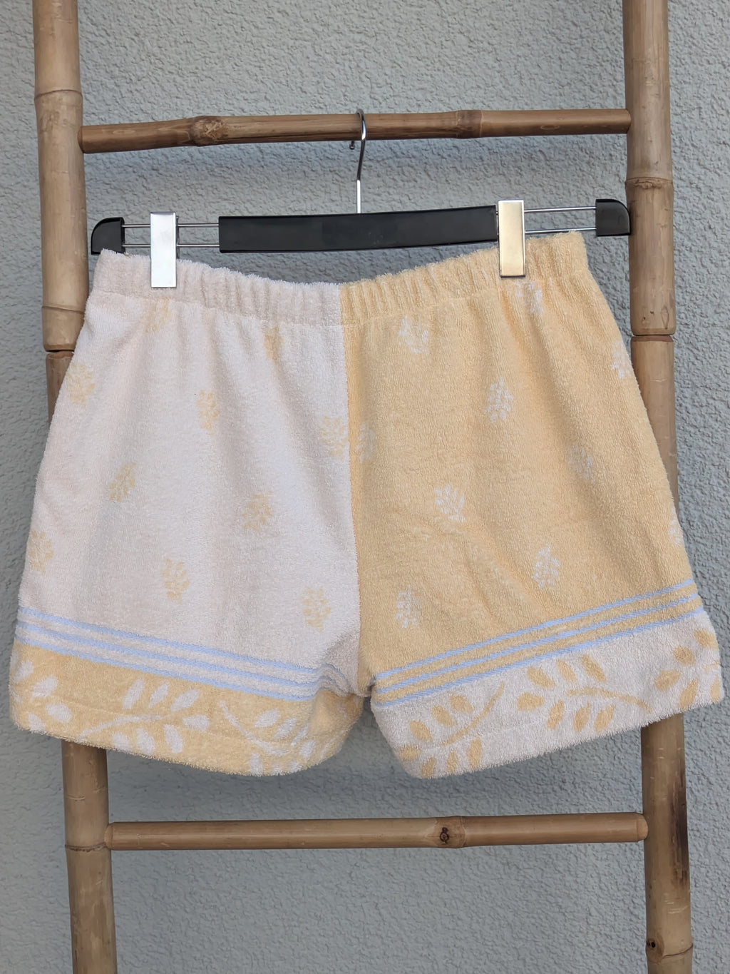 Rückansicht der zweifarbigen Old Wave Towel Shorts 'Buttermilk Breeze '75'. Zeigt die Kombination aus dem pastellgelben und dem cremefarbenen Frotteestoff mit Borte am unteren Saum. Die Hose hängt auf einem Kleiderbügel an einer Bambusleiter.