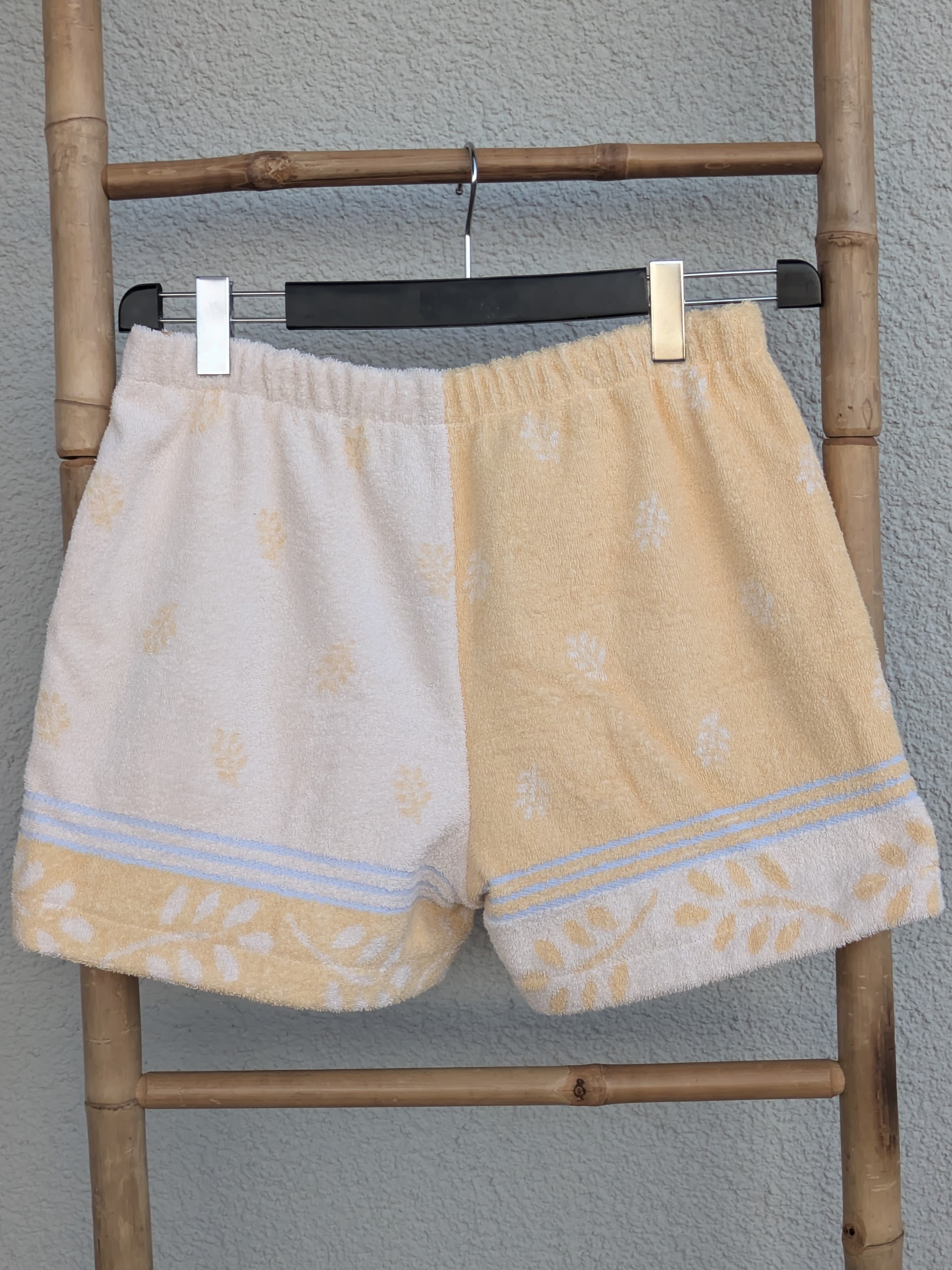 Rückansicht der zweifarbigen Old Wave Towel Shorts 'Buttermilk Breeze '75'. Zeigt die Kombination aus dem pastellgelben und dem cremefarbenen Frotteestoff mit Borte am unteren Saum. Die Hose hängt auf einem Kleiderbügel an einer Bambusleiter.