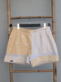 Vorderansicht der Old Wave Towel Shorts 'Buttermilk Breeze '75'. Die linke Hälfte ist cremeweiß mit zartem, pastellgelbem Blumenmuster, die rechte Hälfte ist uni pastellgelb. Die Shorts haben einen hellen elastischen Bund und dekorative Saumbordüren. Das Kleidungsstück hängt an einer Bambusleiter vor einer grauen Wand.