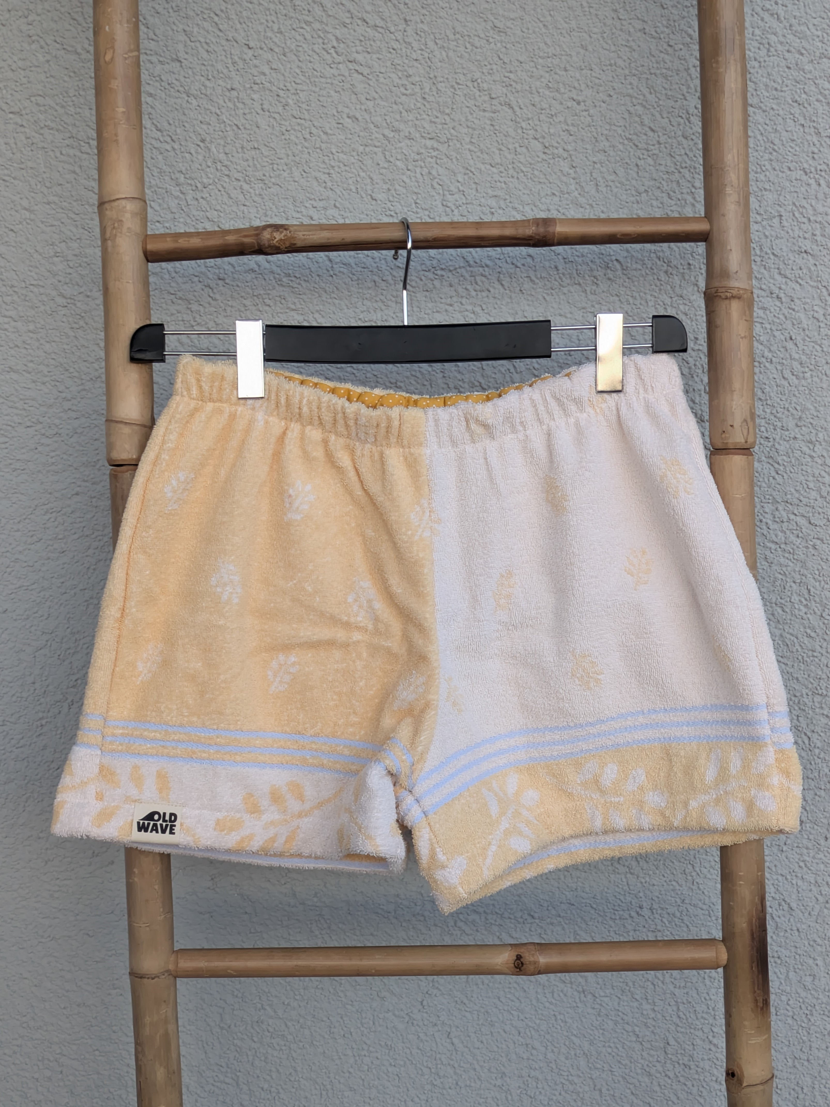 Vorderansicht der Old Wave Towel Shorts 'Buttermilk Breeze '75'. Die linke Hälfte ist cremeweiß mit zartem, pastellgelbem Blumenmuster, die rechte Hälfte ist uni pastellgelb. Die Shorts haben einen hellen elastischen Bund und dekorative Saumbordüren. Das Kleidungsstück hängt an einer Bambusleiter vor einer grauen Wand.