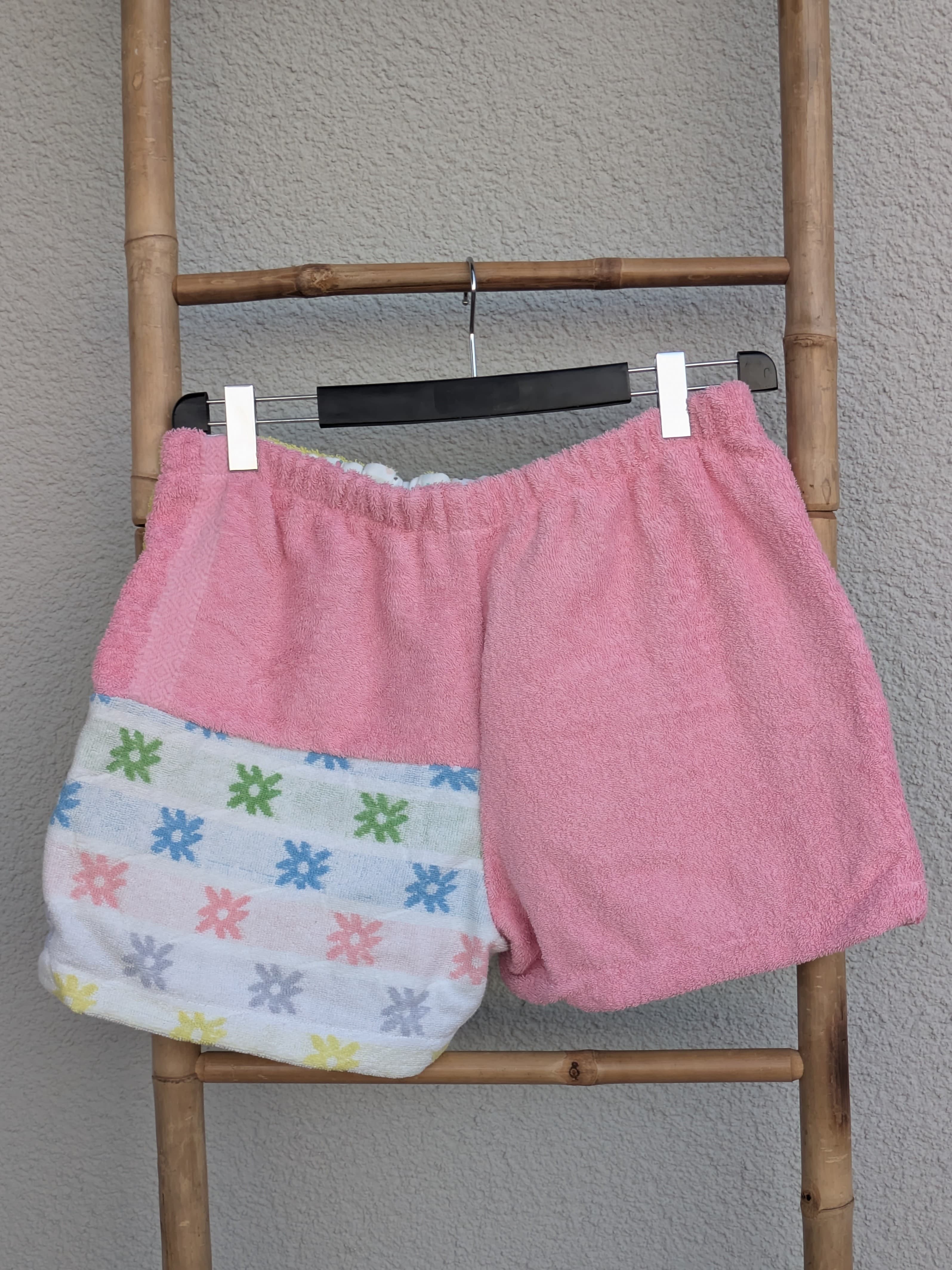 Rückansicht der Frottee-Shorts 'Candy Pop ’77' von Old Wave. Zeigt die vorwiegend rosafarbene Rückseite mit einem bunten Mustereinsatz am linken Hosenbein. Die Shorts hängen an einer Bambusleiter.