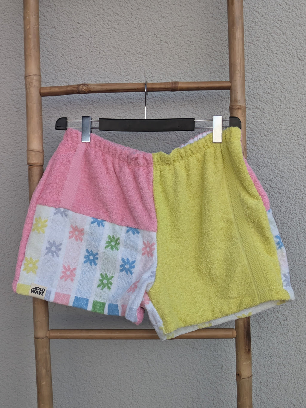 Vorderansicht der bunt gemusterten Old Wave Towel Shorts 'Candy Pop ’77'. Die linke Seite kombiniert Rosa mit einem bunten Vintage-Geometriemuster, die rechte Seite ist gelb. Die Hose hängt an einem Kleiderbügel vor einer hellgrauen Wand.