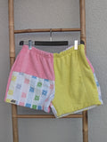 Vorderansicht der bunt gemusterten Old Wave Towel Shorts 'Candy Pop ’77'. Die linke Seite kombiniert Rosa mit einem bunten Vintage-Geometriemuster, die rechte Seite ist gelb. Die Hose hängt an einem Kleiderbügel vor einer hellgrauen Wand.