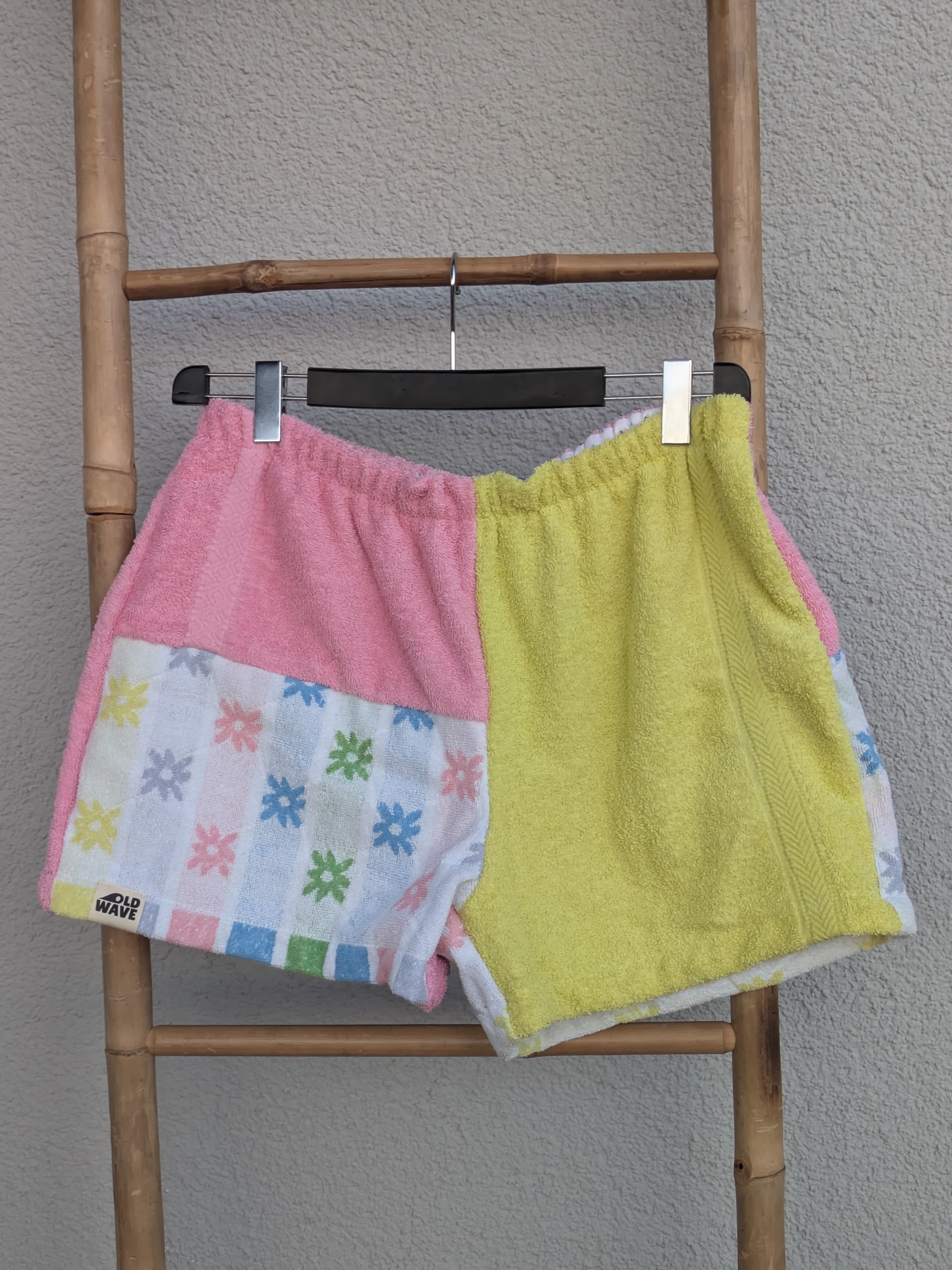 Vorderansicht der bunt gemusterten Old Wave Towel Shorts 'Candy Pop ’77'. Die linke Seite kombiniert Rosa mit einem bunten Vintage-Geometriemuster, die rechte Seite ist gelb. Die Hose hängt an einem Kleiderbügel vor einer hellgrauen Wand.