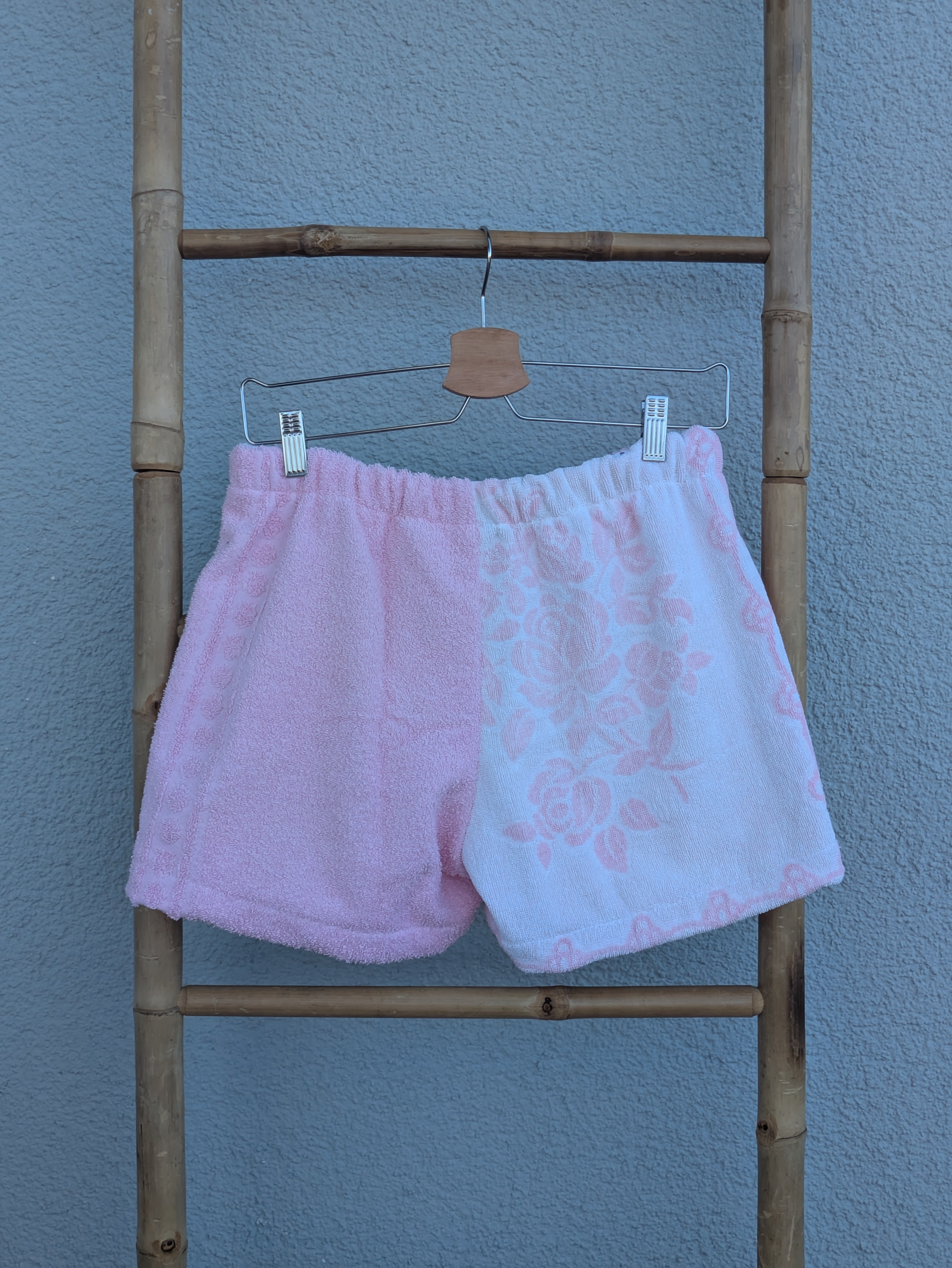 Rückansicht der Frottee Towel Shorts 'Cotton Candy Floral ’71' von Old Wave. Zeigt die zweigeteilte Rückseite in Rosa und weiß-rosa Frottee mit zartem Floral-Muster vor einer hellen Wand.