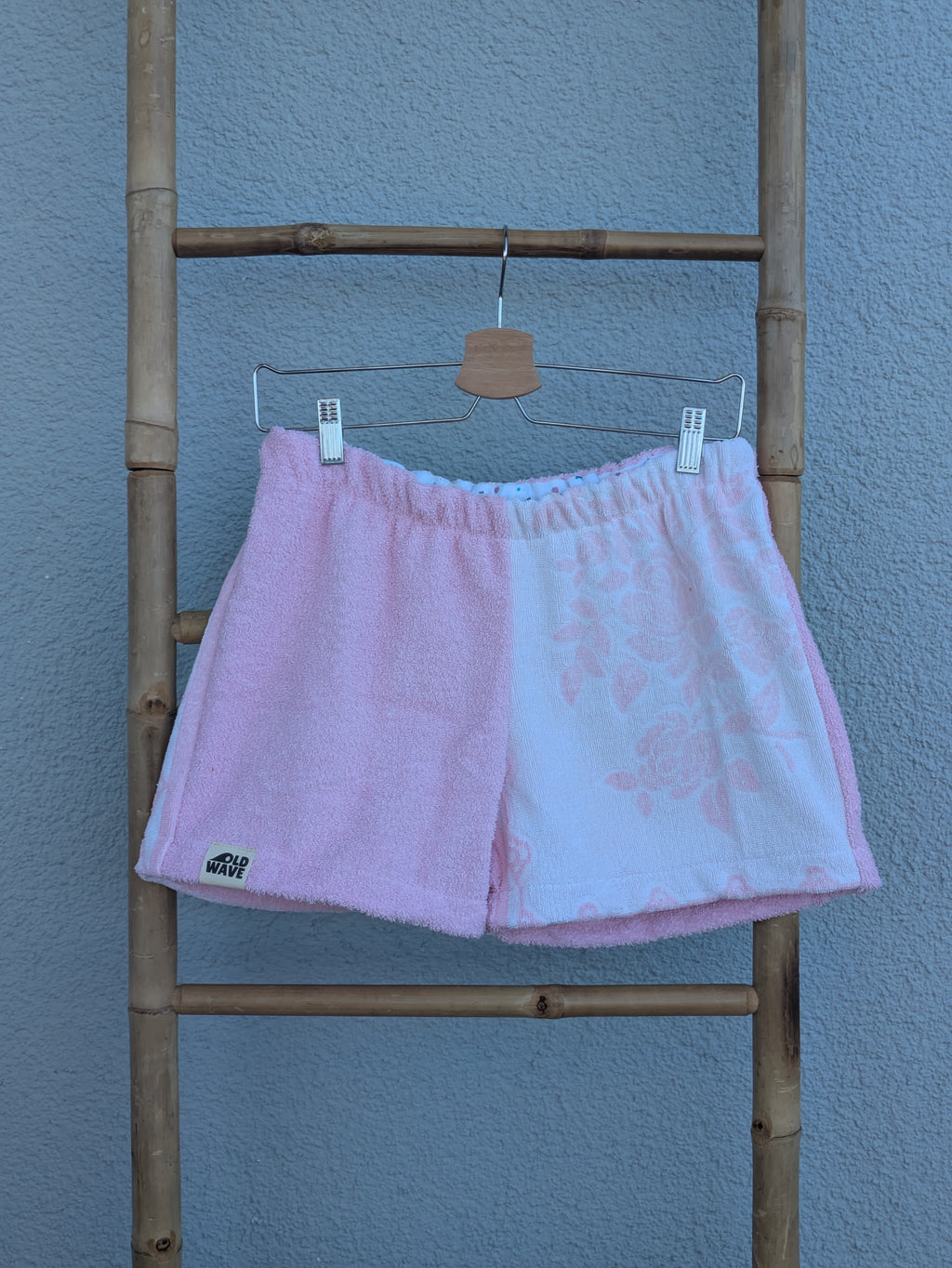 Vorderansicht der Frottee Towel Shorts 'Cotton Candy Floral ’71' von Old Wave in Zartrosa und weißem Musterfrottee. Zweigeteiltes Two-Tone-Design mit unifarbener rosa Seite und zart eingewebtem Blumenmotiv auf der hellen Hosenhälfte, präsentiert an einer Bambusleiter.