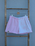 Vorderansicht der Frottee Towel Shorts 'Cotton Candy Floral ’71' von Old Wave in Zartrosa und weißem Musterfrottee. Zweigeteiltes Two-Tone-Design mit unifarbener rosa Seite und zart eingewebtem Blumenmotiv auf der hellen Hosenhälfte, präsentiert an einer Bambusleiter.