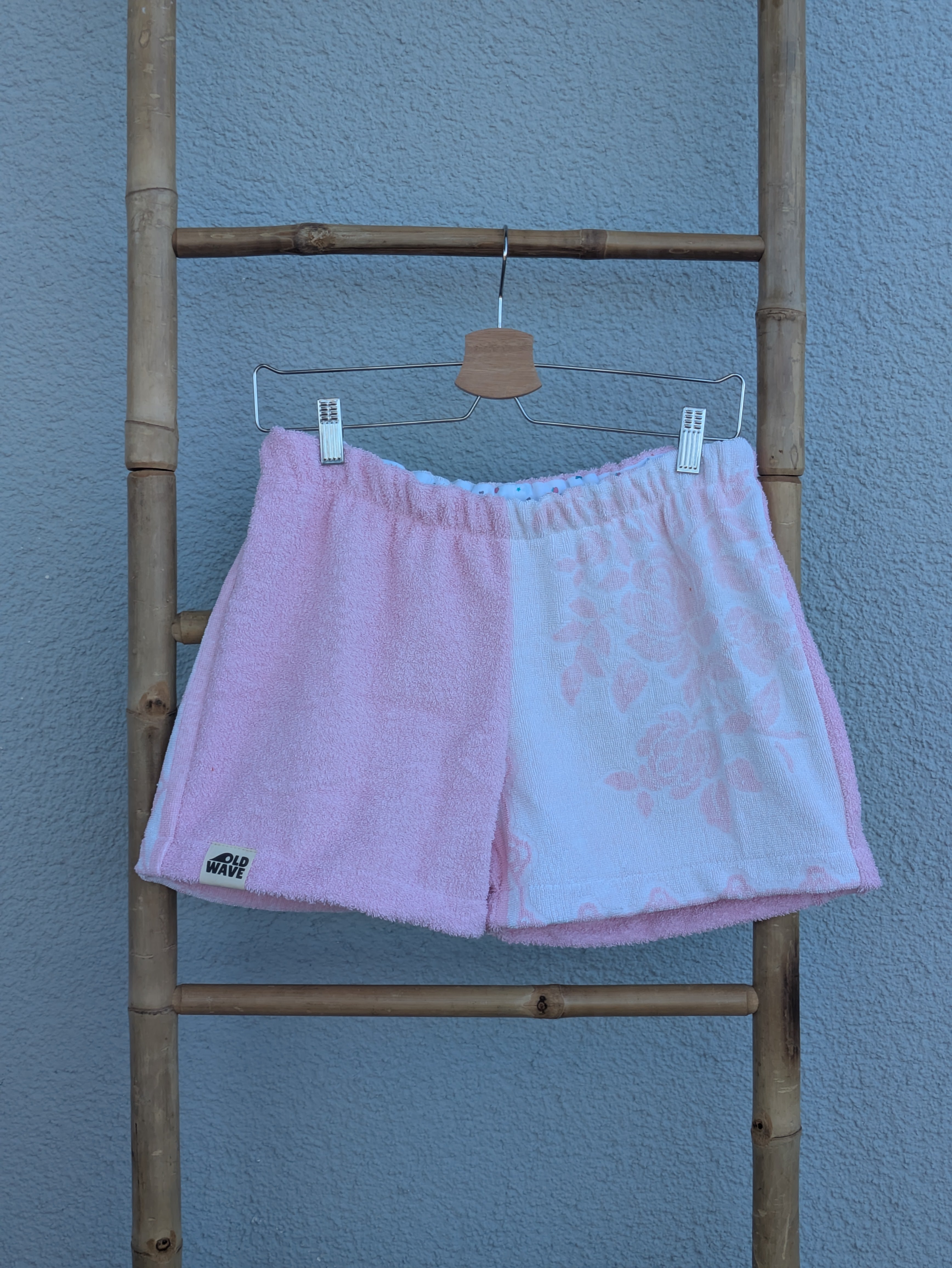 Vorderansicht der Frottee Towel Shorts 'Cotton Candy Floral ’71' von Old Wave in Zartrosa und weißem Musterfrottee. Zweigeteiltes Two-Tone-Design mit unifarbener rosa Seite und zart eingewebtem Blumenmotiv auf der hellen Hosenhälfte, präsentiert an einer Bambusleiter.