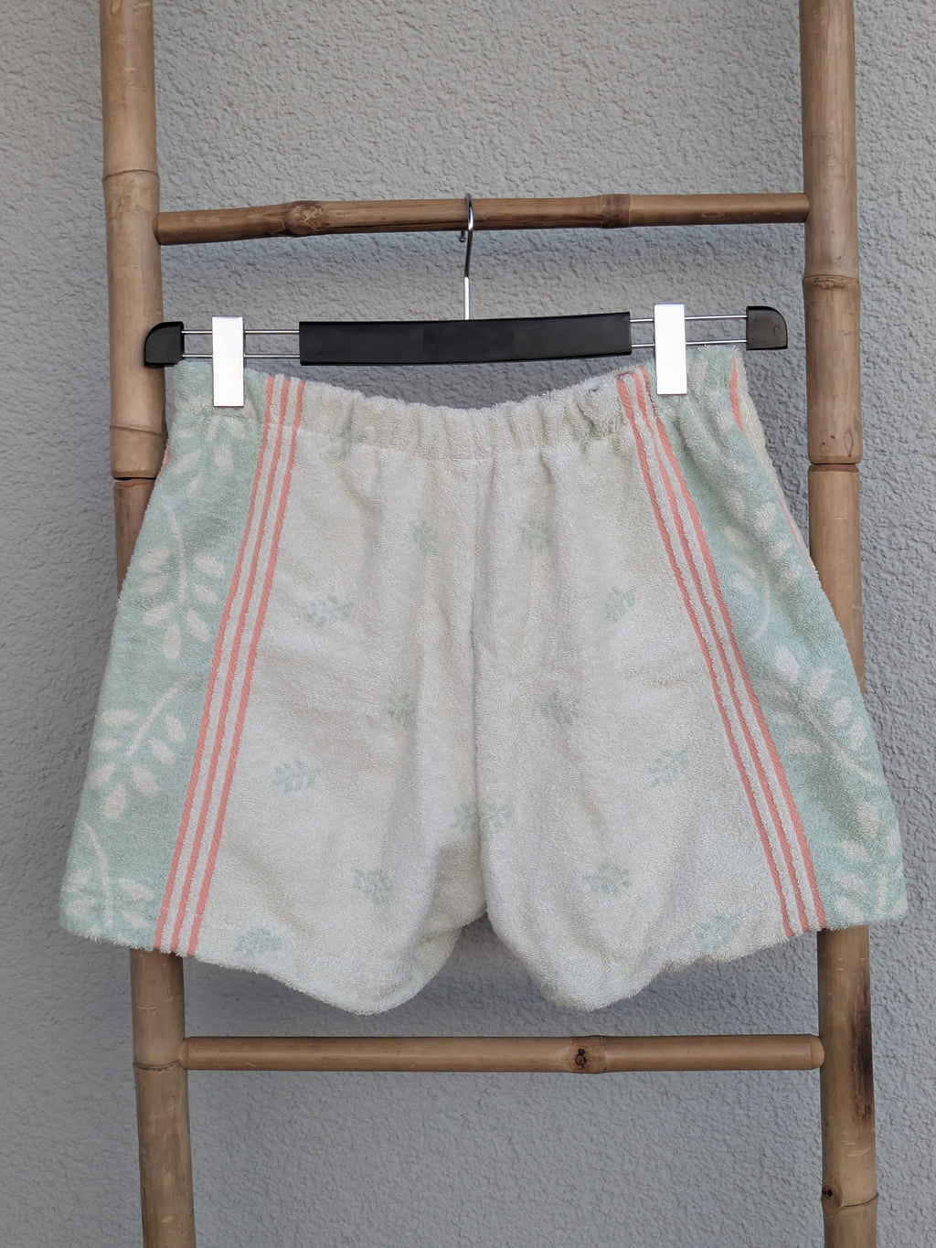 Rückansicht der Frottee-Shorts 'Eucalyptus Breeze ’70' von Old Wave. Zeigt den hellen Hauptstoff sowie die seitlich verlaufenden mintgrünen und rosa Details. Die Hose hängt an einer Bambusleiter.