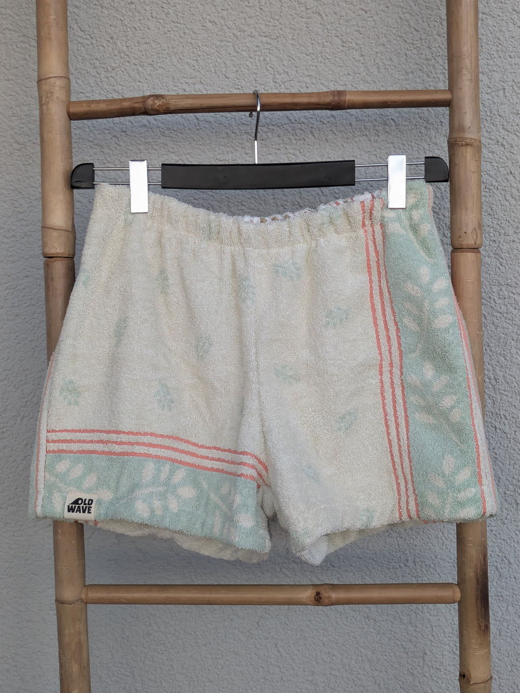 Vorderansicht der Old Wave Towel Shorts 'Eucalyptus Breeze ’70'. Cremeweiße Frottee-Shorts mit seitlichen mintgrünen Mustereinsätzen, abgegrenzt durch zartrosa Paspelstreifen. Die Hose hängt an einem Kleiderbügel vor einer hellen Wand.