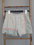 Vorderansicht der Old Wave Towel Shorts 'Eucalyptus Breeze ’70'. Cremeweiße Frottee-Shorts mit seitlichen mintgrünen Mustereinsätzen, abgegrenzt durch zartrosa Paspelstreifen. Die Hose hängt an einem Kleiderbügel vor einer hellen Wand.