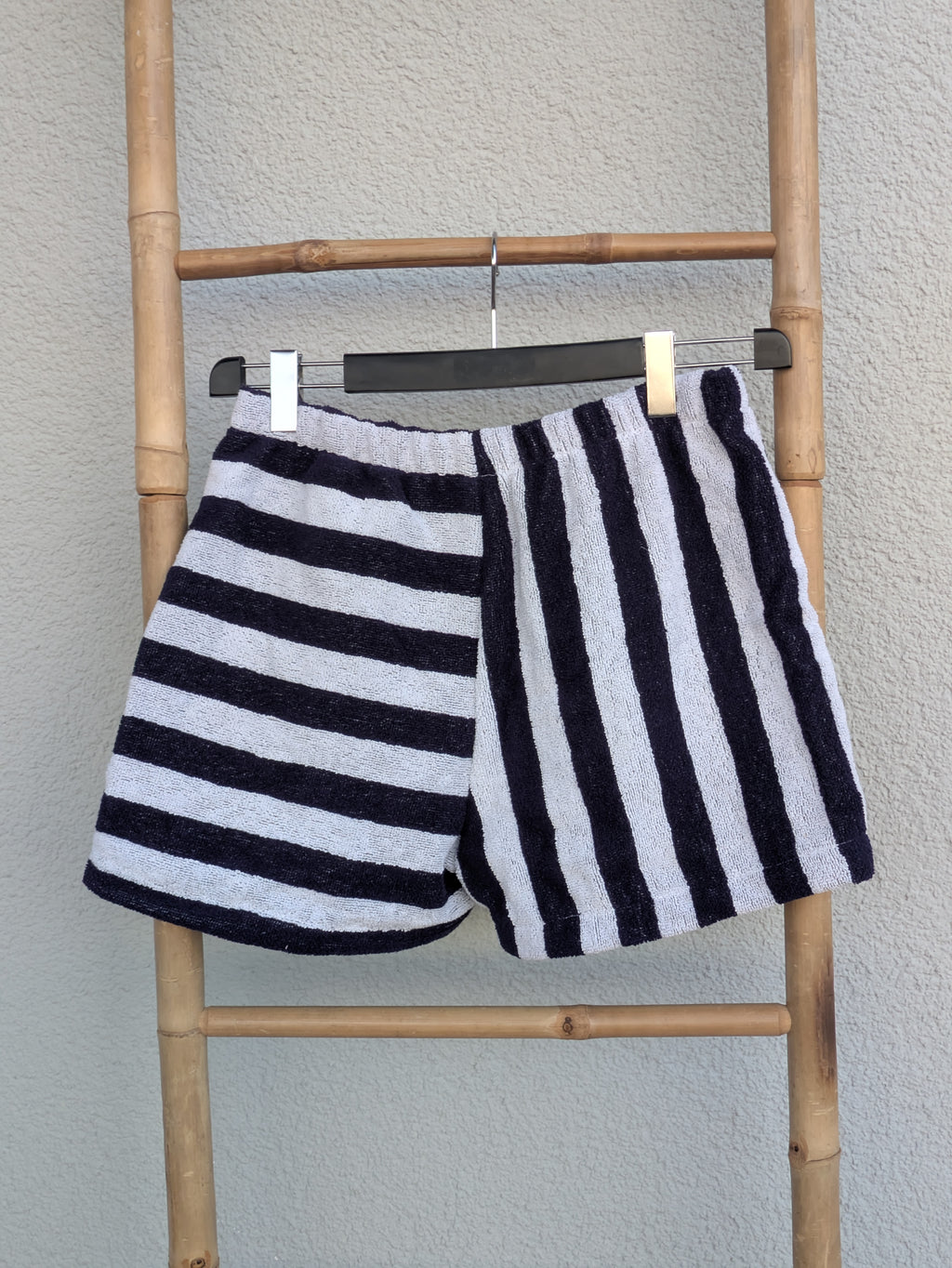 Rückansicht der zweifarbigen gestreiften Frottee-Shorts 'Nautical Wave ’78' von Old Wave. Zeigt das Zusammenspiel aus waagerechten und senkrechten blau-weißen Streifen. Die Shorts hängen an einer Bambusleiter.