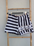 Vorderansicht der Old Wave Towel Shorts 'Nautical Wave ’78'. Die linke Seite hat waagerechte blau-weiße Streifen, die rechte Seite vertikale Streifen mit einer dekorativen Anker-Bordüre am unteren Saum. Die Hose hängt an einem Kleiderbügel vor einer hellen Wand.