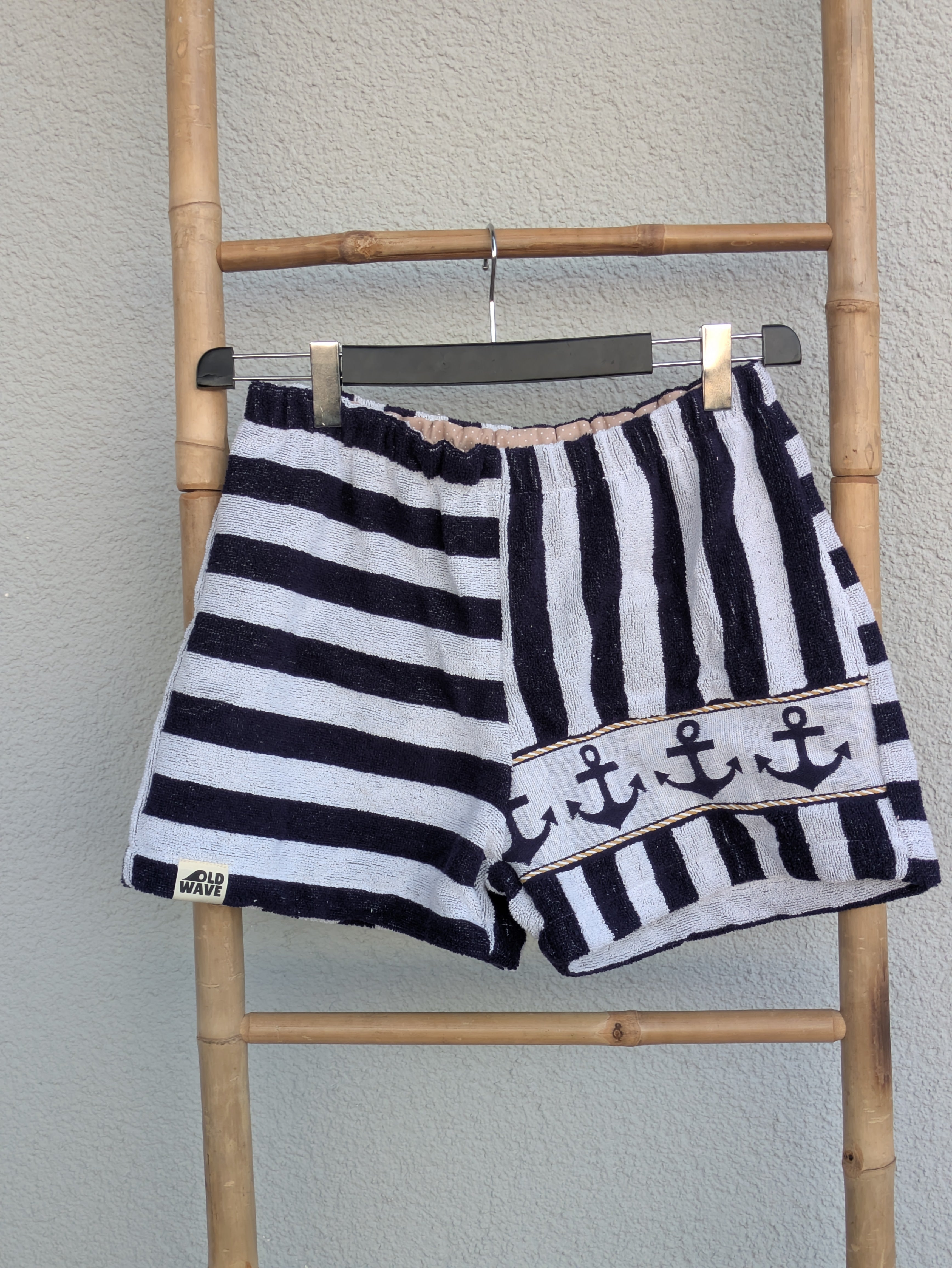 Vorderansicht der Old Wave Towel Shorts 'Nautical Wave ’78'. Die linke Seite hat waagerechte blau-weiße Streifen, die rechte Seite vertikale Streifen mit einer dekorativen Anker-Bordüre am unteren Saum. Die Hose hängt an einem Kleiderbügel vor einer hellen Wand.