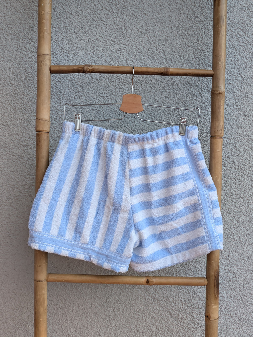 Rückansicht der Frottee Towel Shorts 'Ocean Heart Stripe ’74' von Old Wave. Zeigt das Streifen-Design in Hellblau und Weiß mit längs gestreiftem Hosenbein links und quer gestreifter rechter Hosenhälfte vor einer hellen Wand.