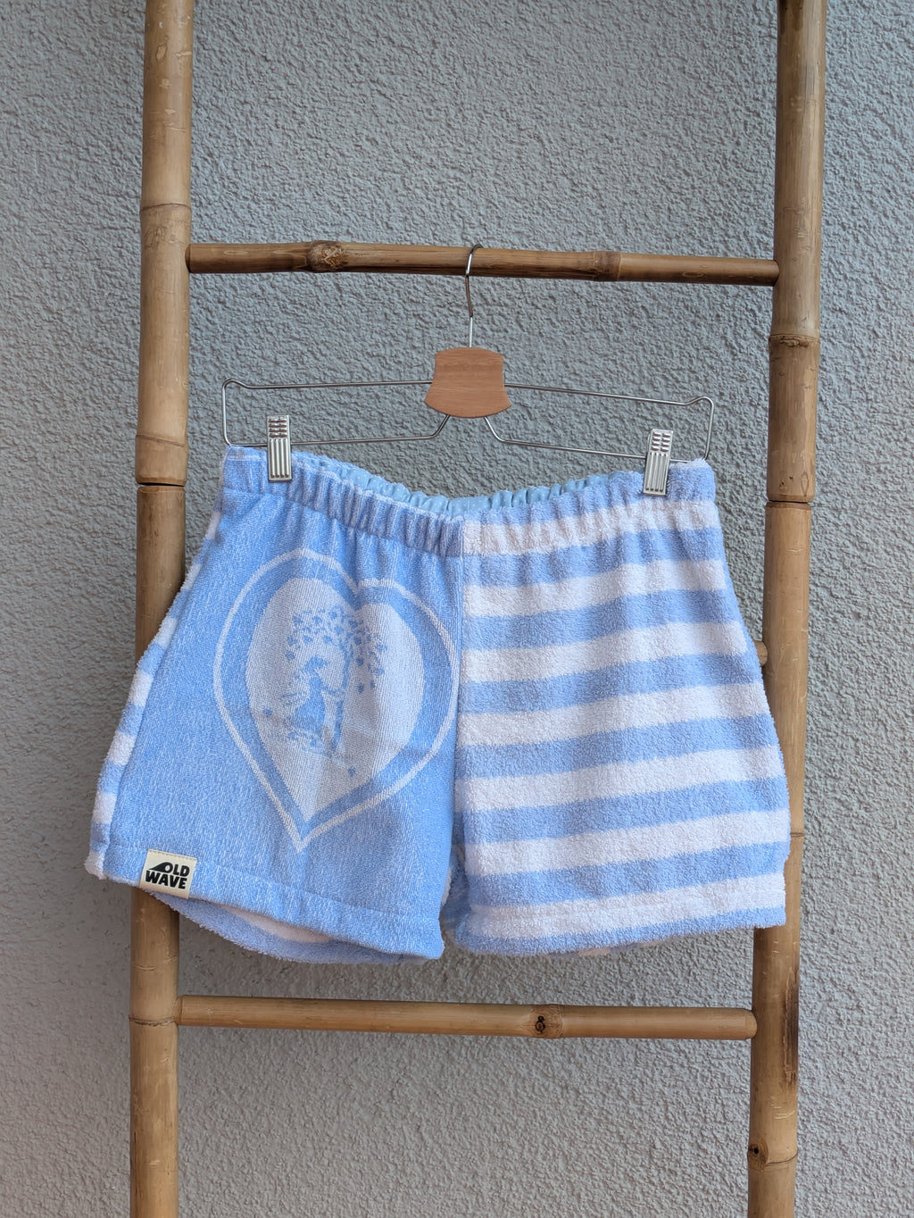 Vorderansicht der Frottee Towel Shorts 'Ocean Heart Stripe ’74' von Old Wave in Hellblau und Weiß. Zweigeteiltes Two-Tone-Design mit großem eingewebten Herzmotiv links, quer gestreiftem Frotteemuster rechts und Logo-Patch unten links, präsentiert an einer Bambusleiter.