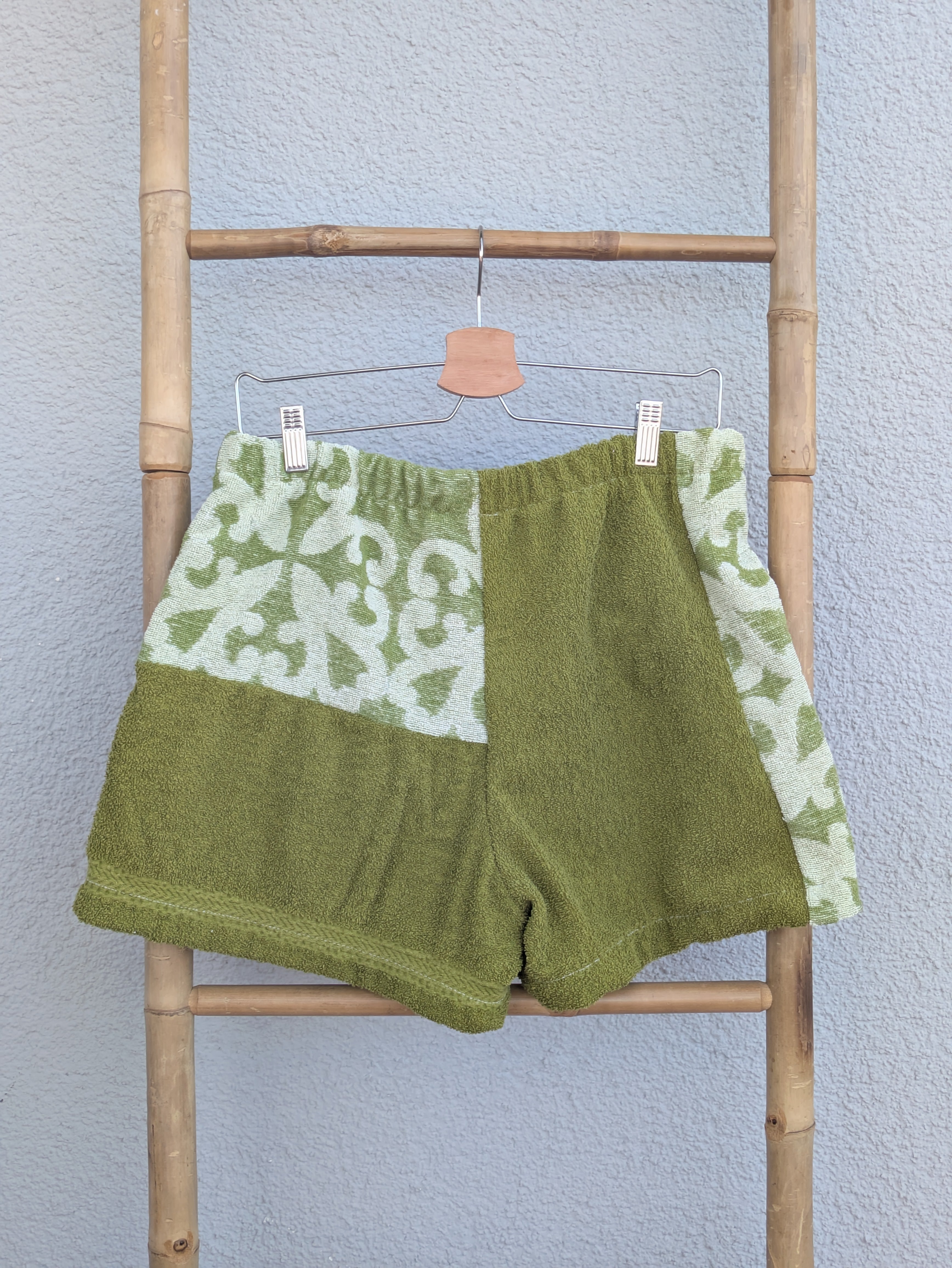 Rückansicht der Frottee Towel Shorts 'Olive Ornament ’75' von Old Wave. Zeigt die Rückseite in unifarbenem Olivgrün mit gemustertem Passen-Einsatz oben links und gemustertem Seiteneinsatz rechts vor einer hellen Wand.